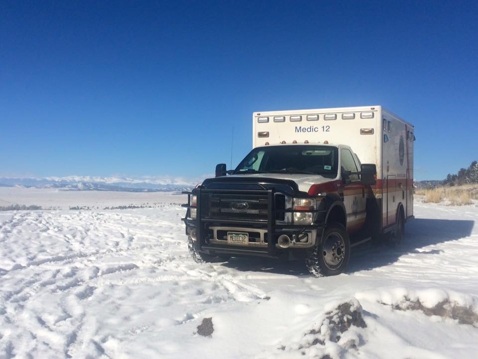 An emergency medical vehicle parked in a snowy landscape under a clear blue sky, with mountains in the distance.
