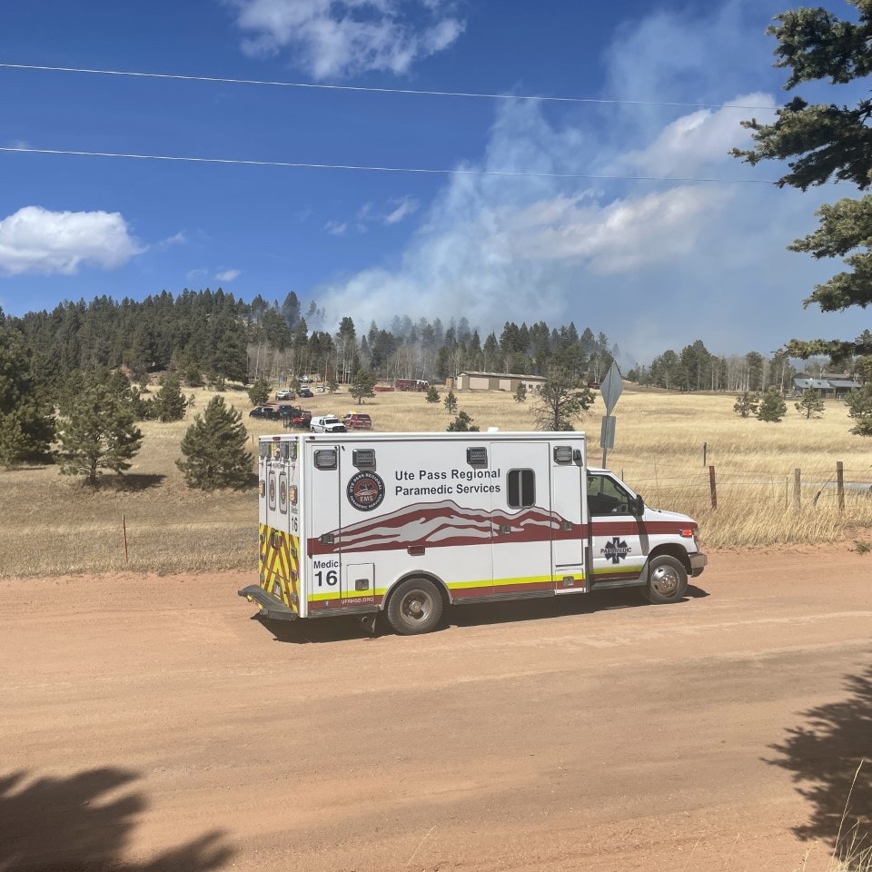 An ambulance is parked on a dirt road with smoke rising from a nearby area, possibly indicating a fire. Emergency response is visible.
