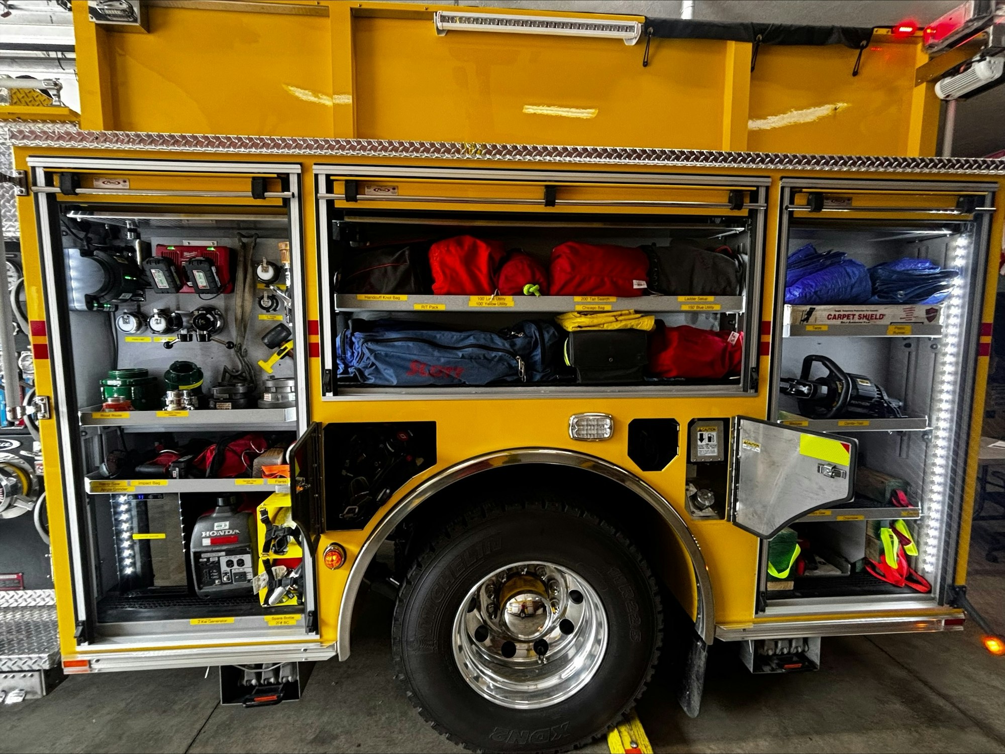 The image shows the inside of a fire truck compartment, equipped with various tools, gear, and supplies organized on shelves.