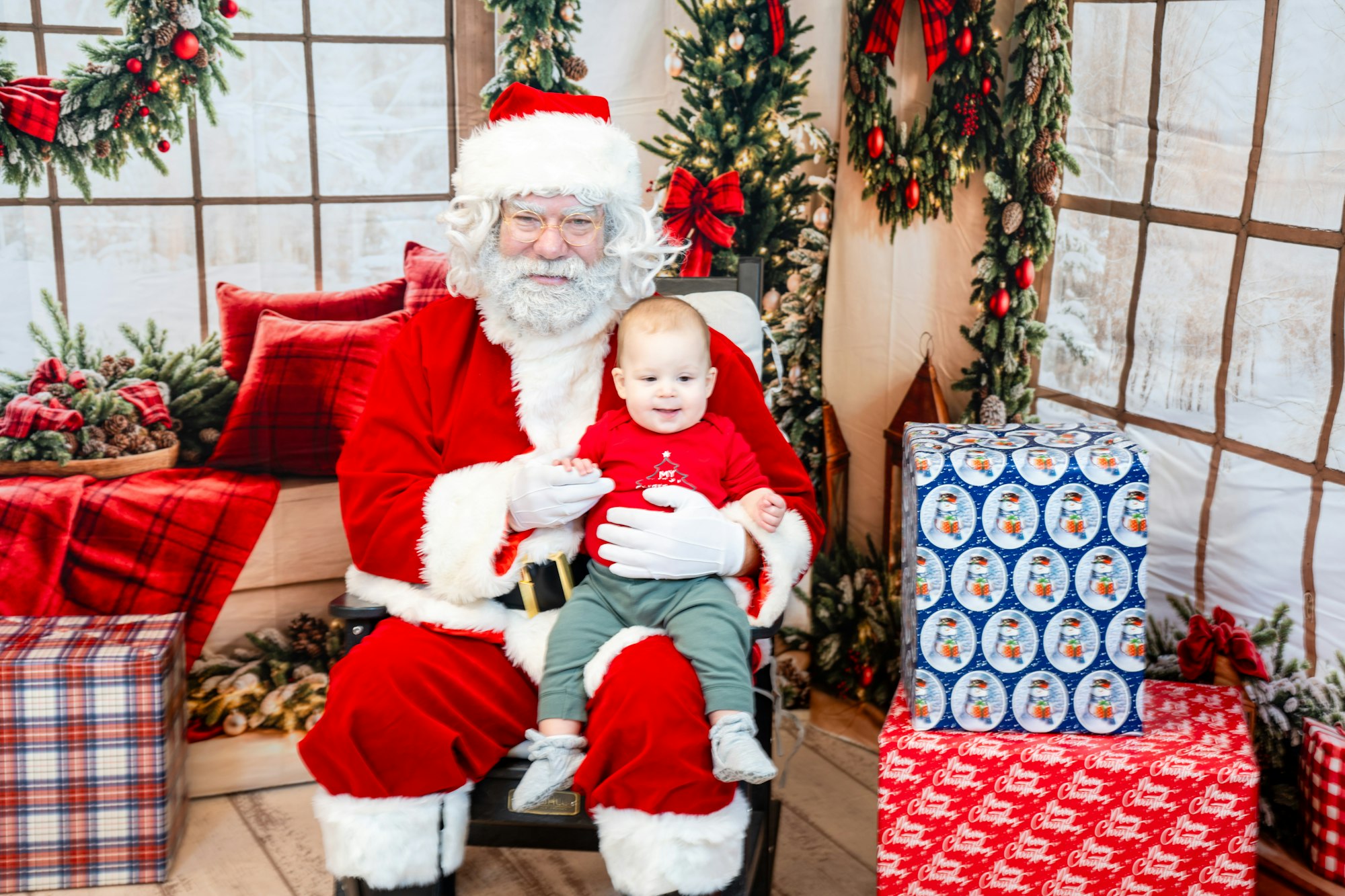 A Santa Claus holding a baby, surrounded by festive decorations and wrapped gifts in a cozy holiday setting.