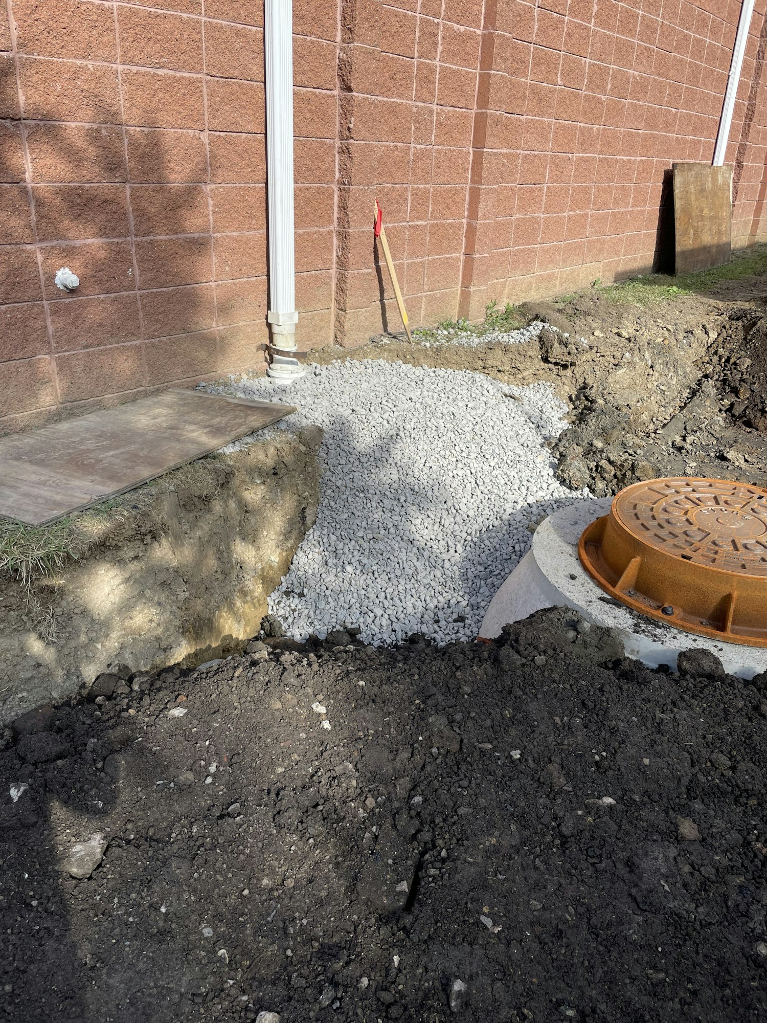 Excavated area with gravel by a manhole cover, next to tiled pavement and grass, with a shovel visible.