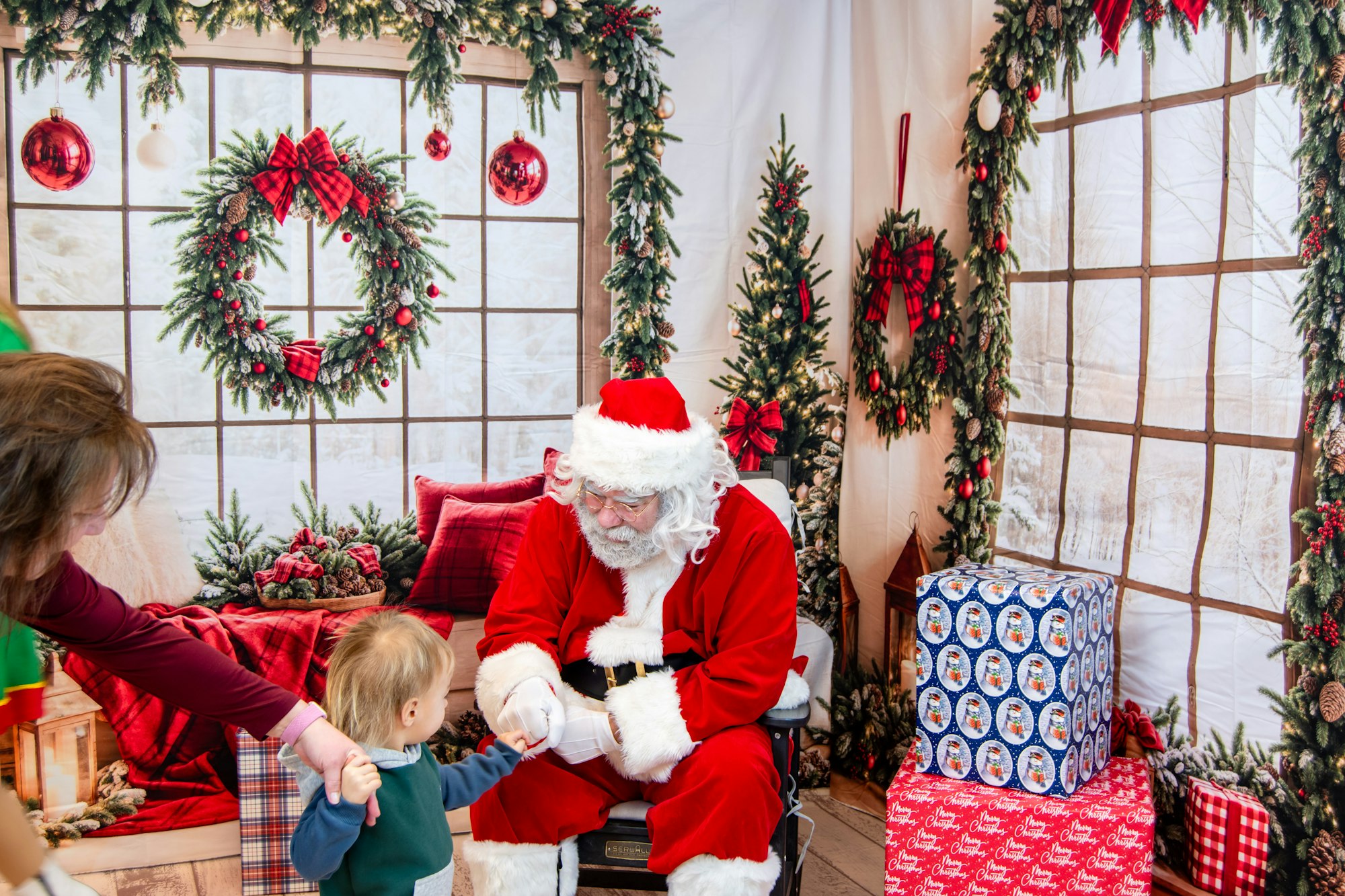 A cheerful Santa interacts with a young child in a festive holiday setting, surrounded by decorations and gift boxes.