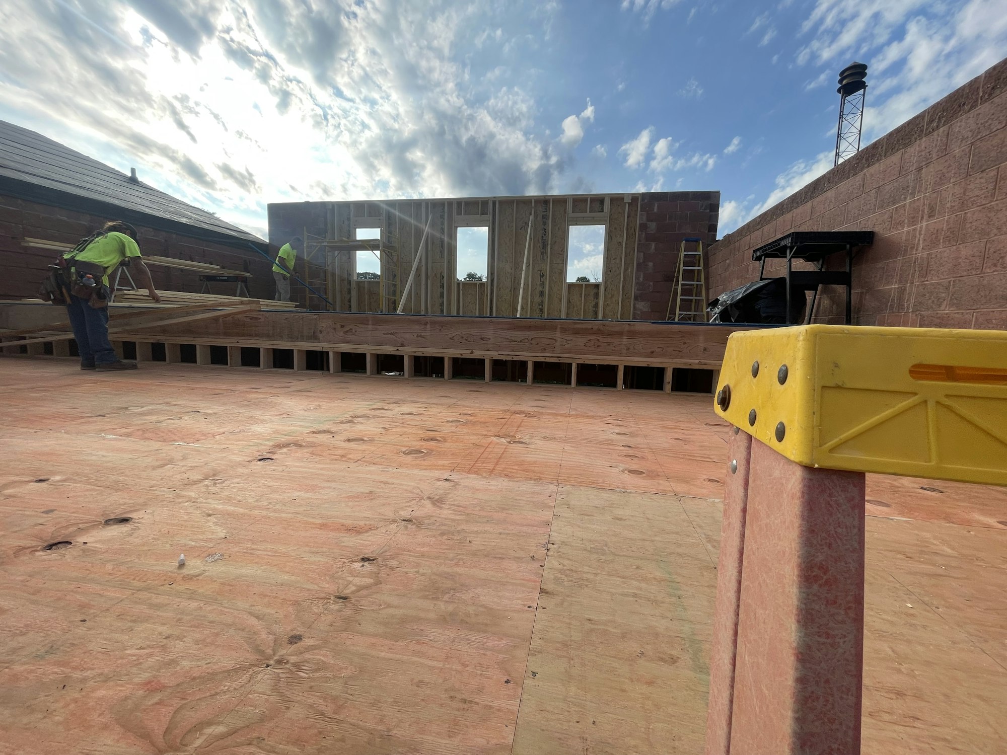 Construction site with workers, wooden framing, and brick walls under a cloudy sky.