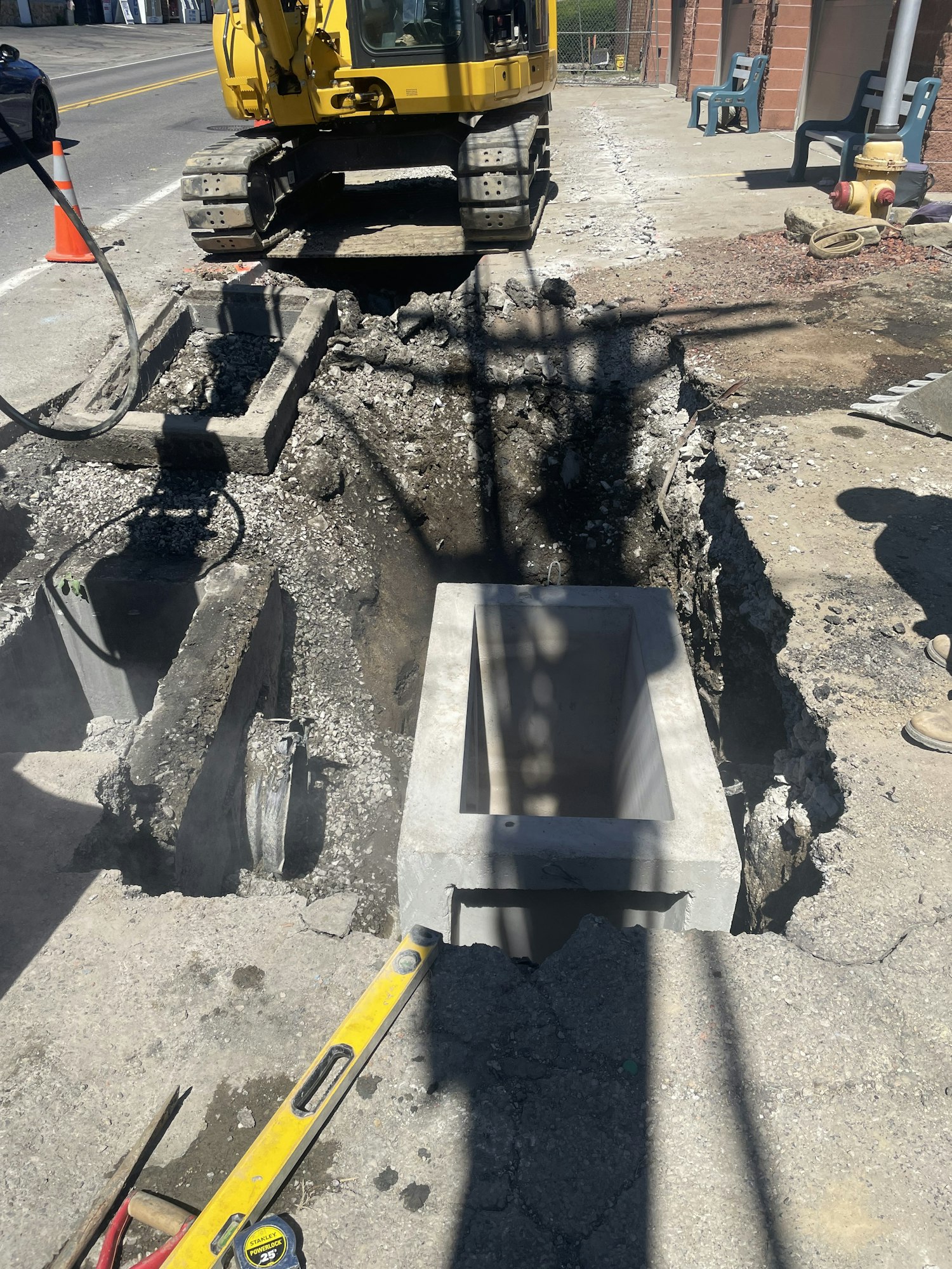 A road under repair with a large hole, construction machinery, a level tool, and debris visible. Image is rotated.