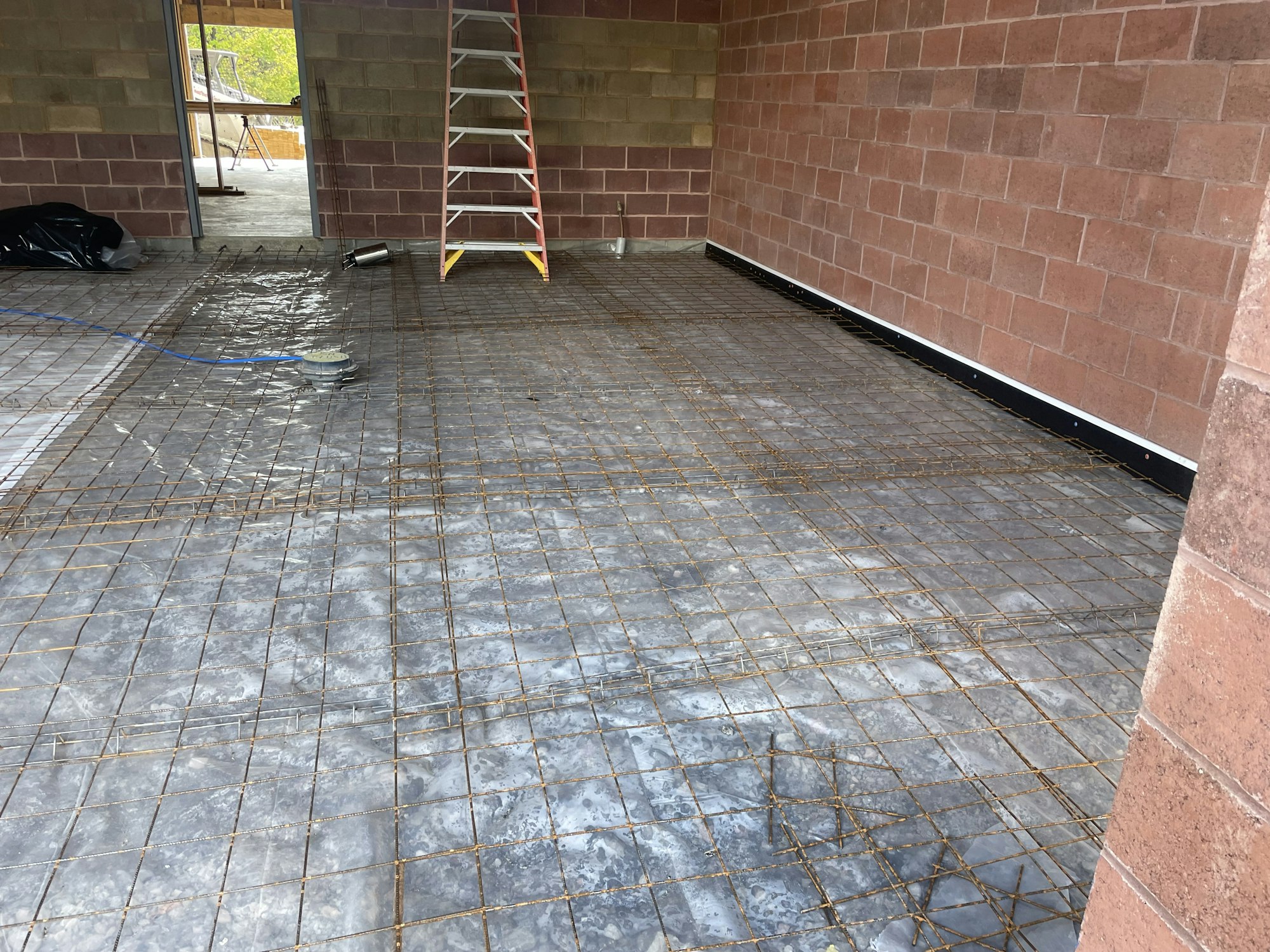 Construction site with metal rebar grid on floor prepping for concrete pour, ladder, and plastic sheeting.