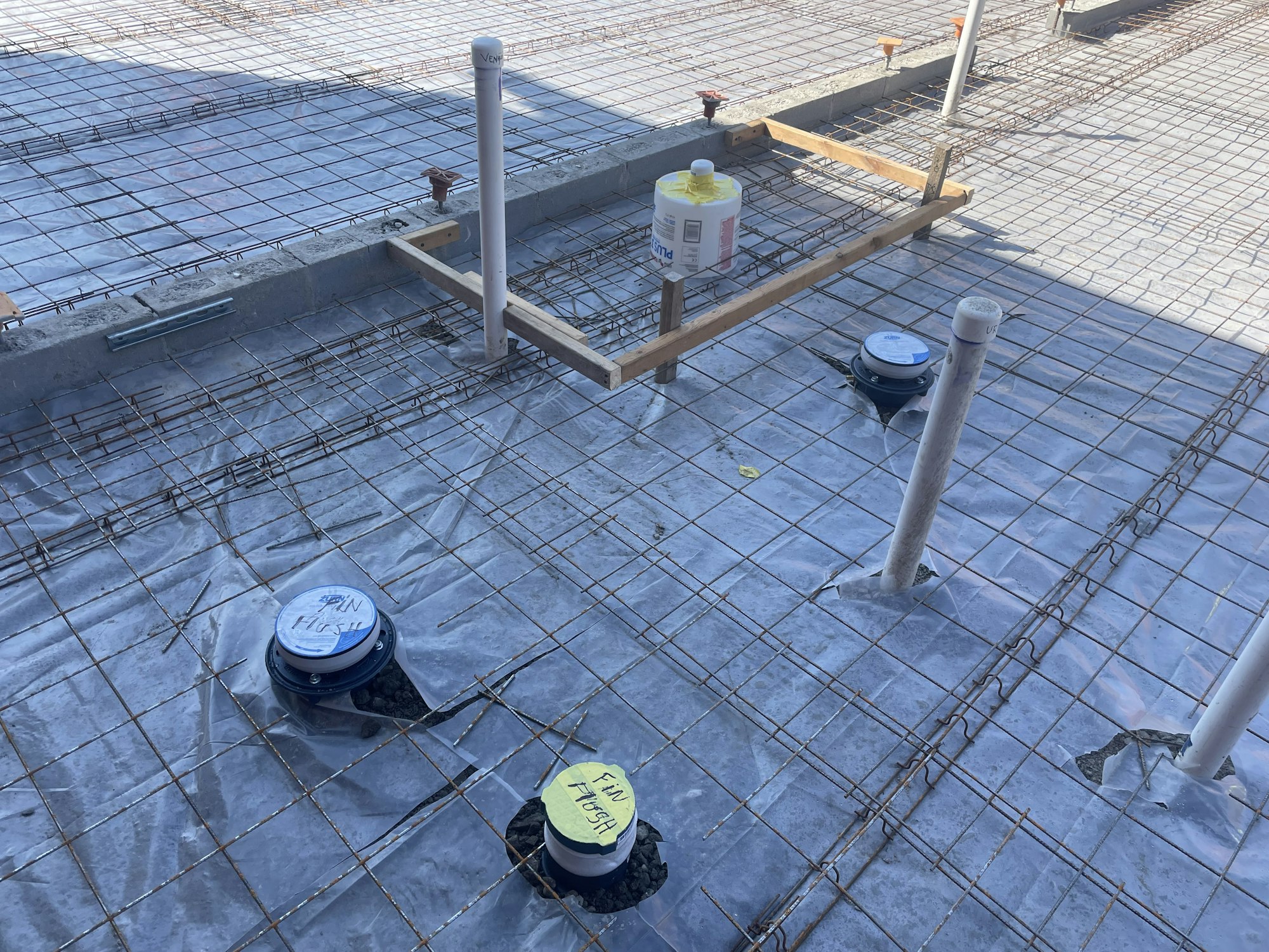 A construction site with rebar grid, pipes, and utility access points before concrete pour.