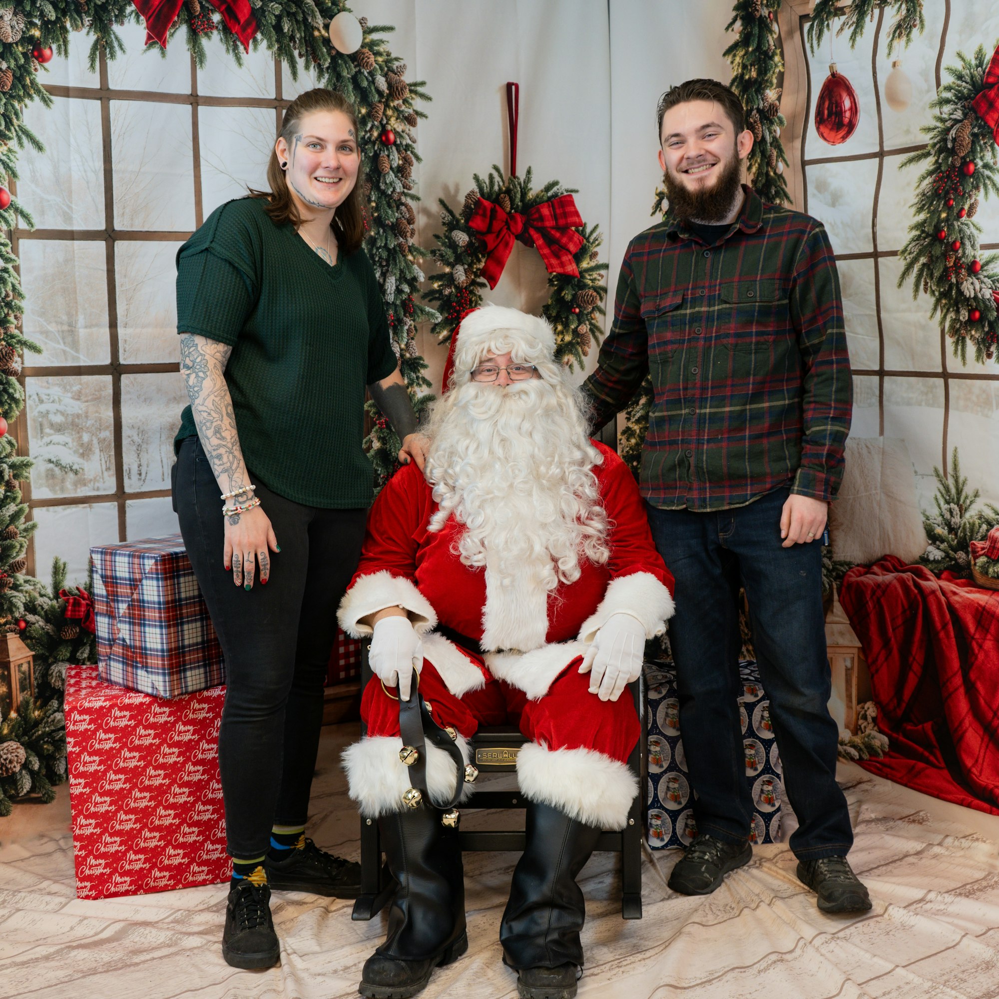 Two people pose with Santa Claus in a festive, decorated setting with wrapped gifts.