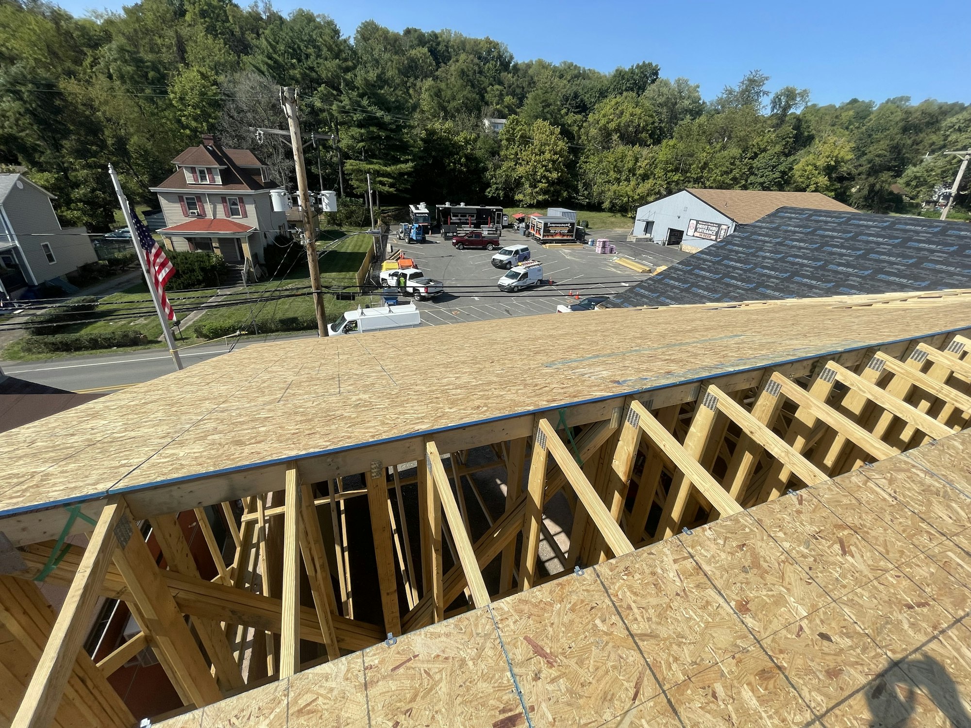 Construction site with wooden framing, an American flag, residential area, vehicles, and trees in the background.