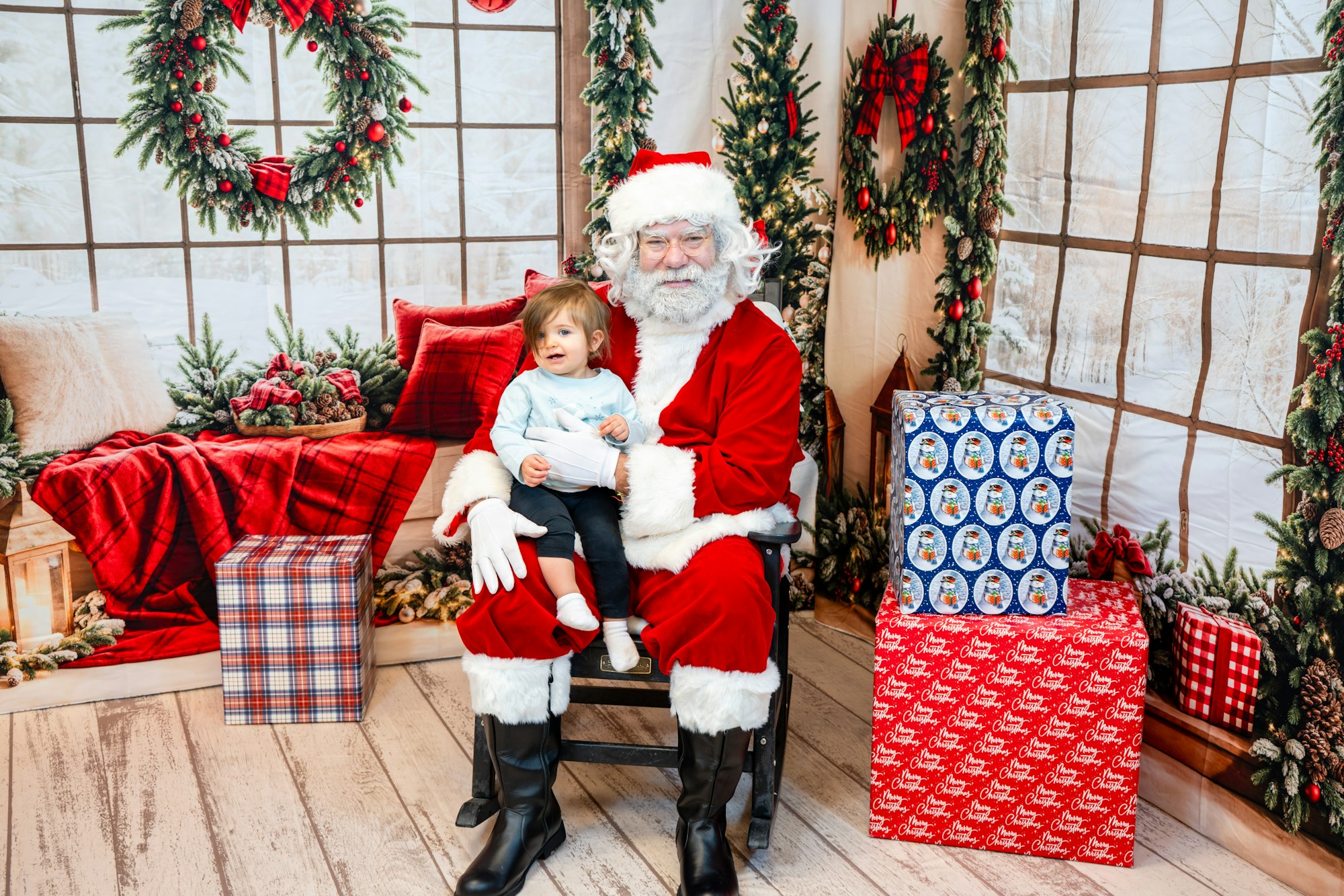 A child sits on Santa's lap in a festive setting with holiday decorations and wrapped gifts around them.