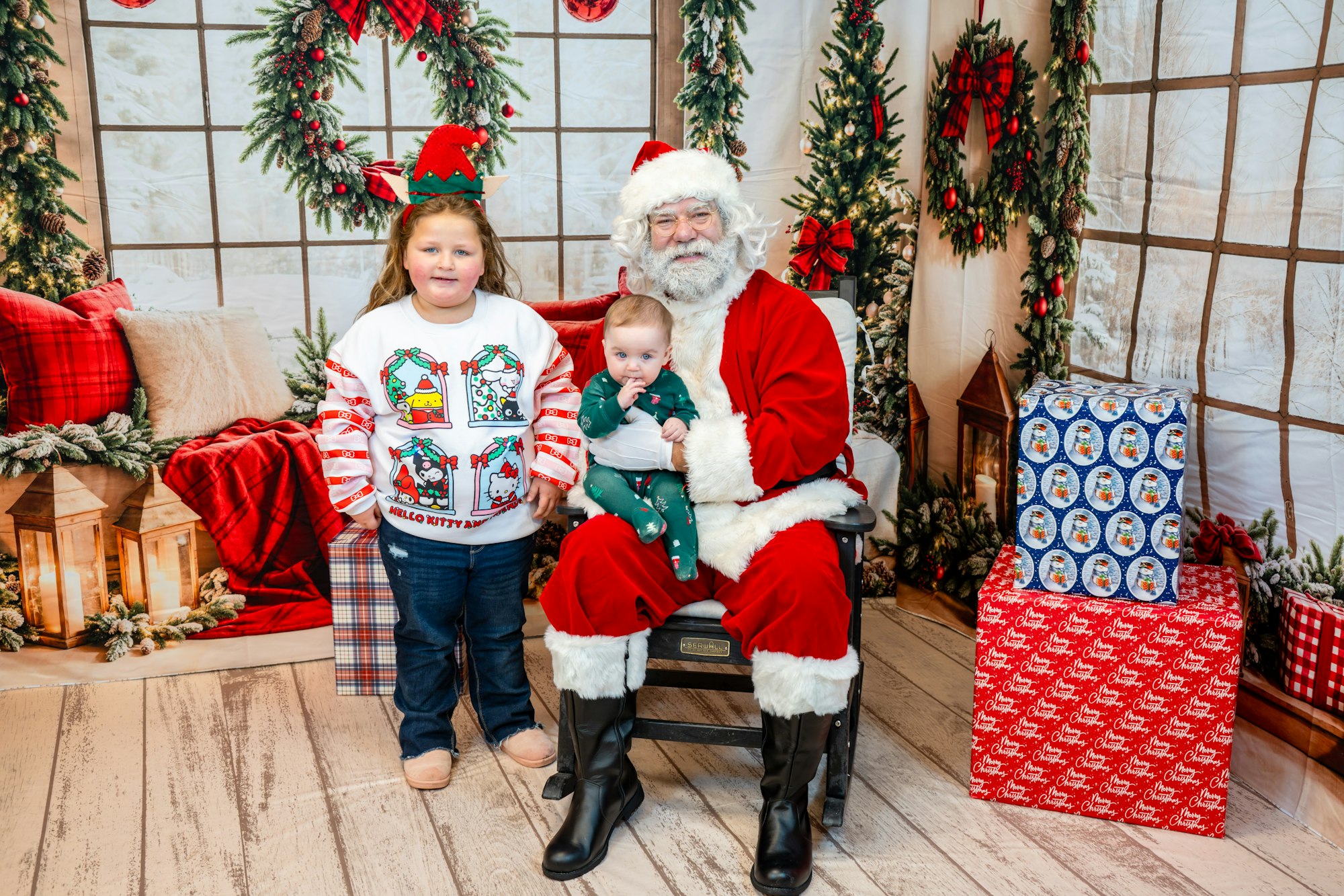 A child in a festive sweater and an infant sit with Santa Claus, surrounded by holiday decorations and wrapped gifts.