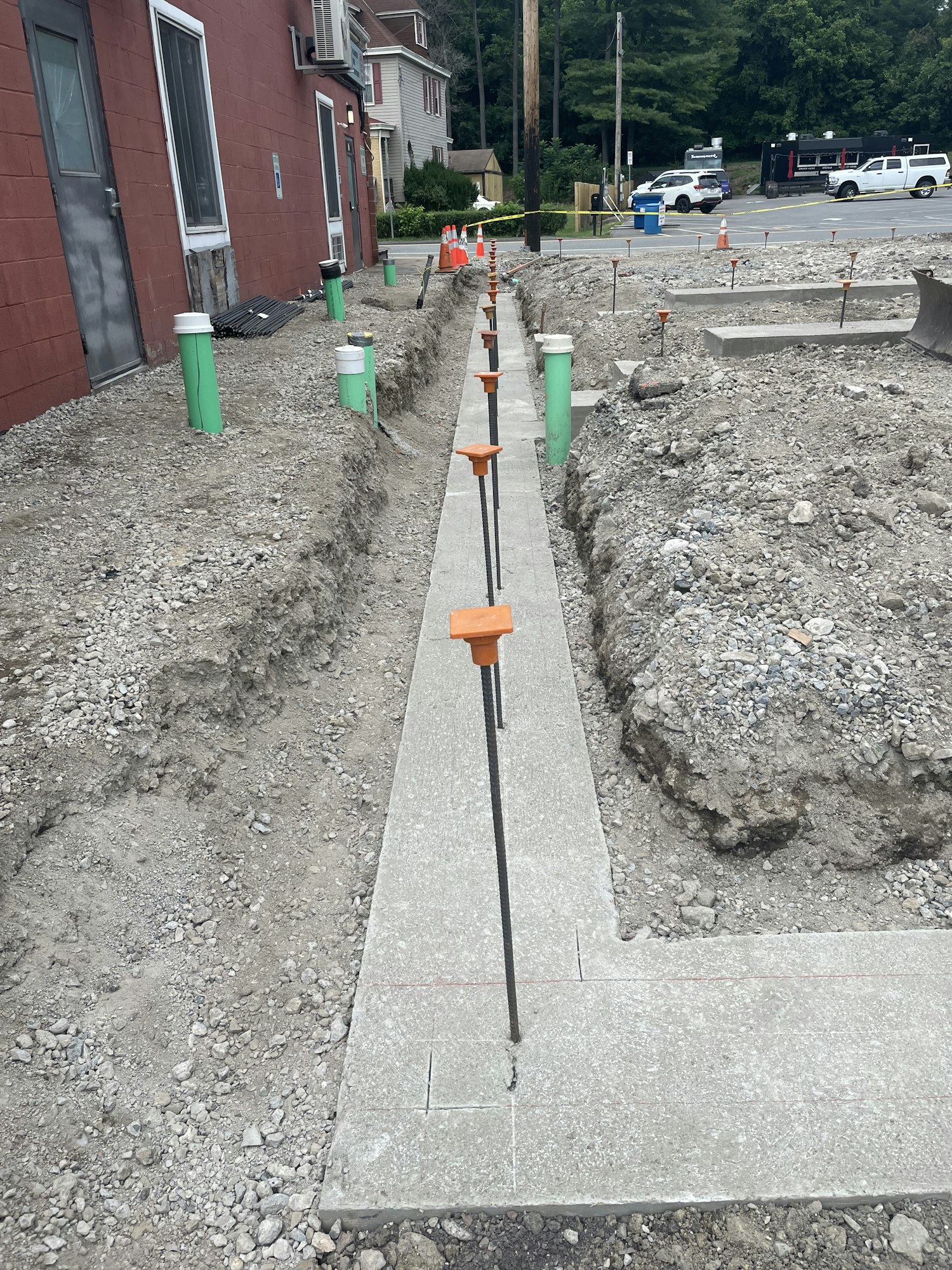 A construction site with concrete slabs, pipes protruding from the ground, traffic cones, and a vehicle in the background.