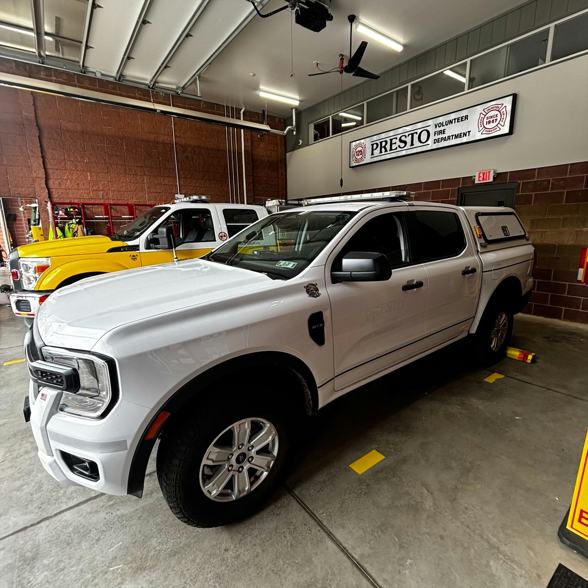 A white truck is parked inside a fire station with a "Presto Volunteer Fire Department" sign, alongside a yellow vehicle.