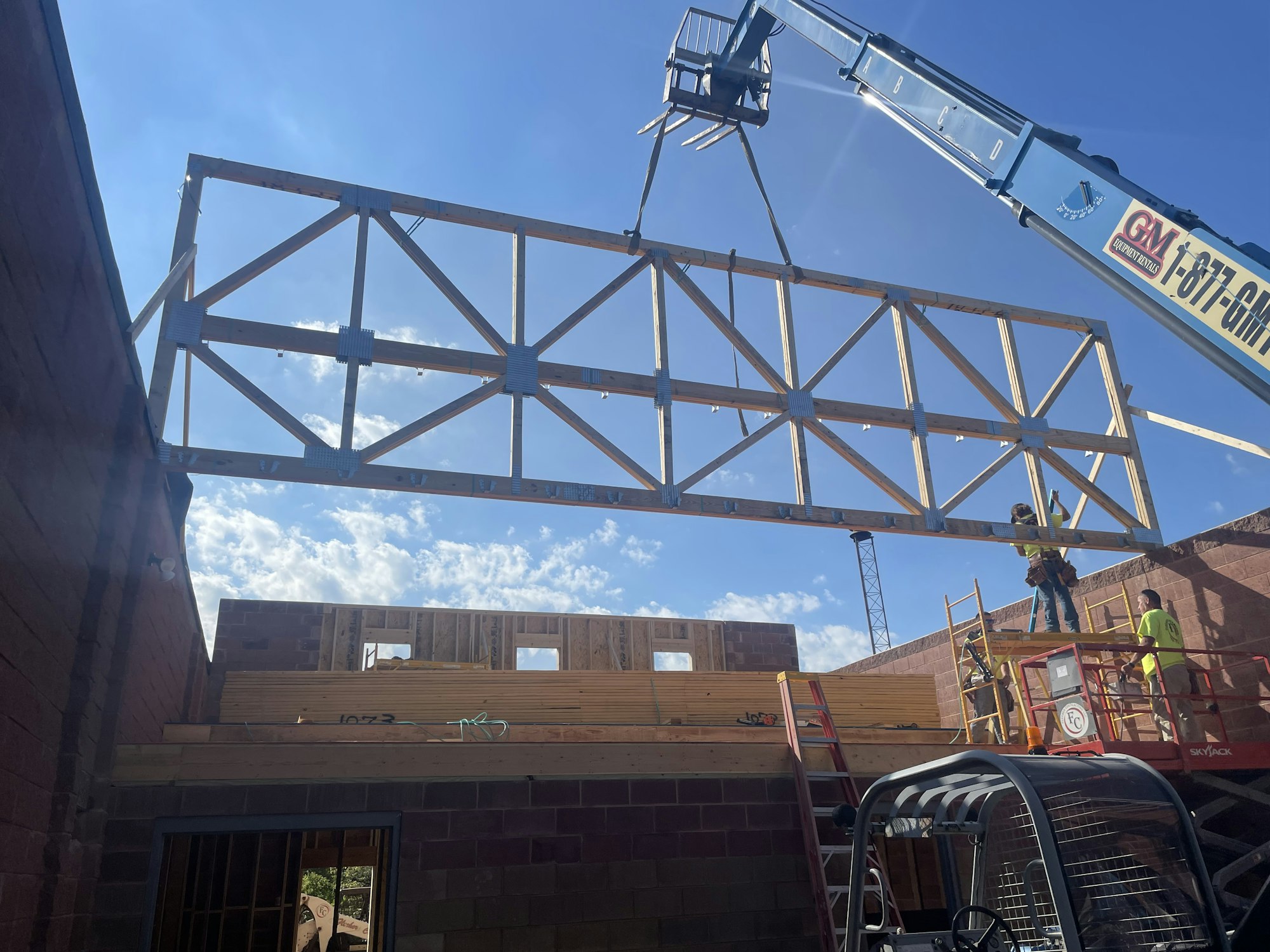 A large metal truss being lifted by a crane onto a building under construction with workers assisting.