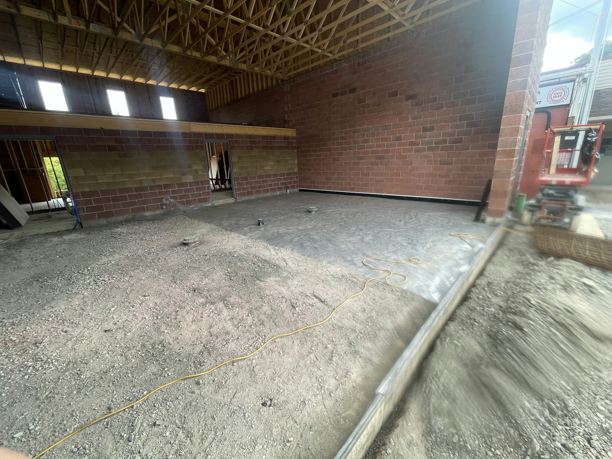 An unfinished interior space with exposed brick walls, a gravel floor, wooden trusses, and construction equipment.
