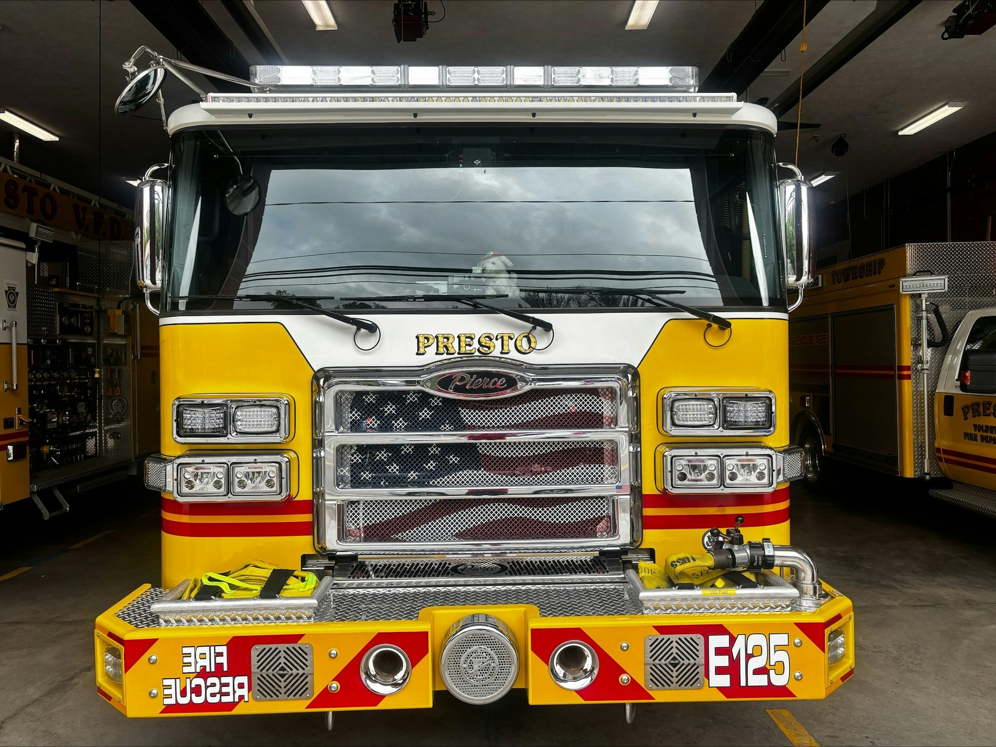 A bright yellow fire truck with "PRESTO" on the front, featuring emergency lights and a rescue hose at the station.