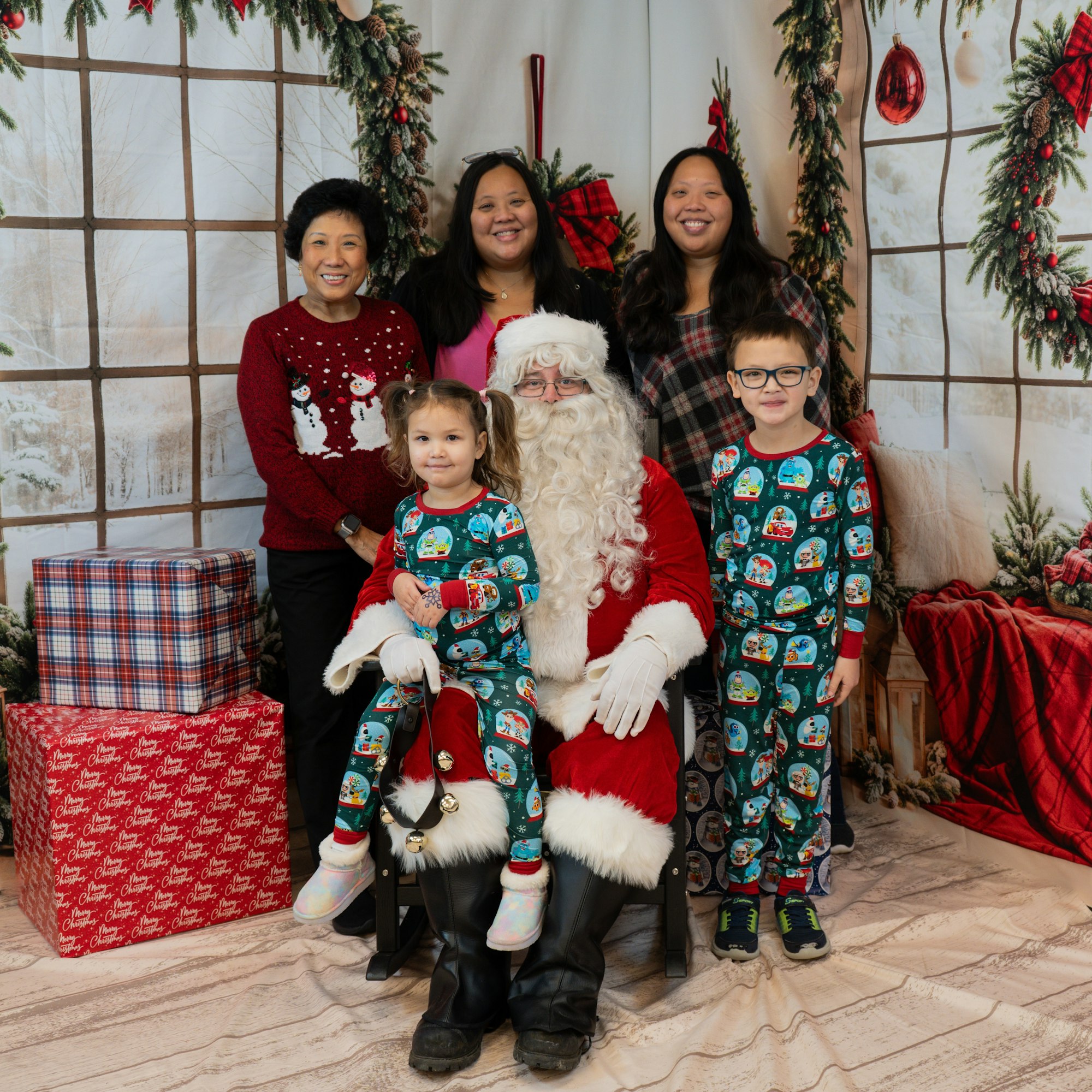 A family poses with Santa, surrounded by Christmas decorations and gifts. Kids wear matching pajamas.
