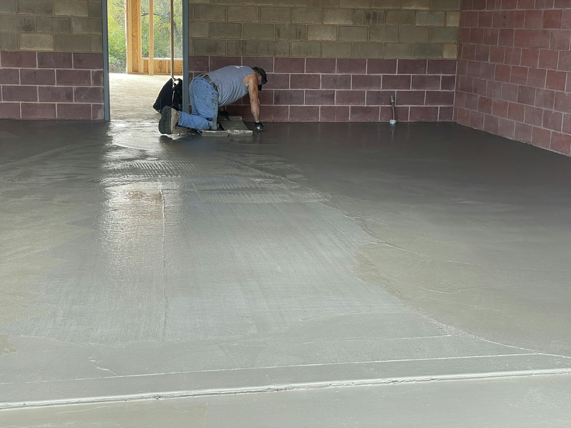 Worker smoothing wet concrete floor with trowel in a room under construction.