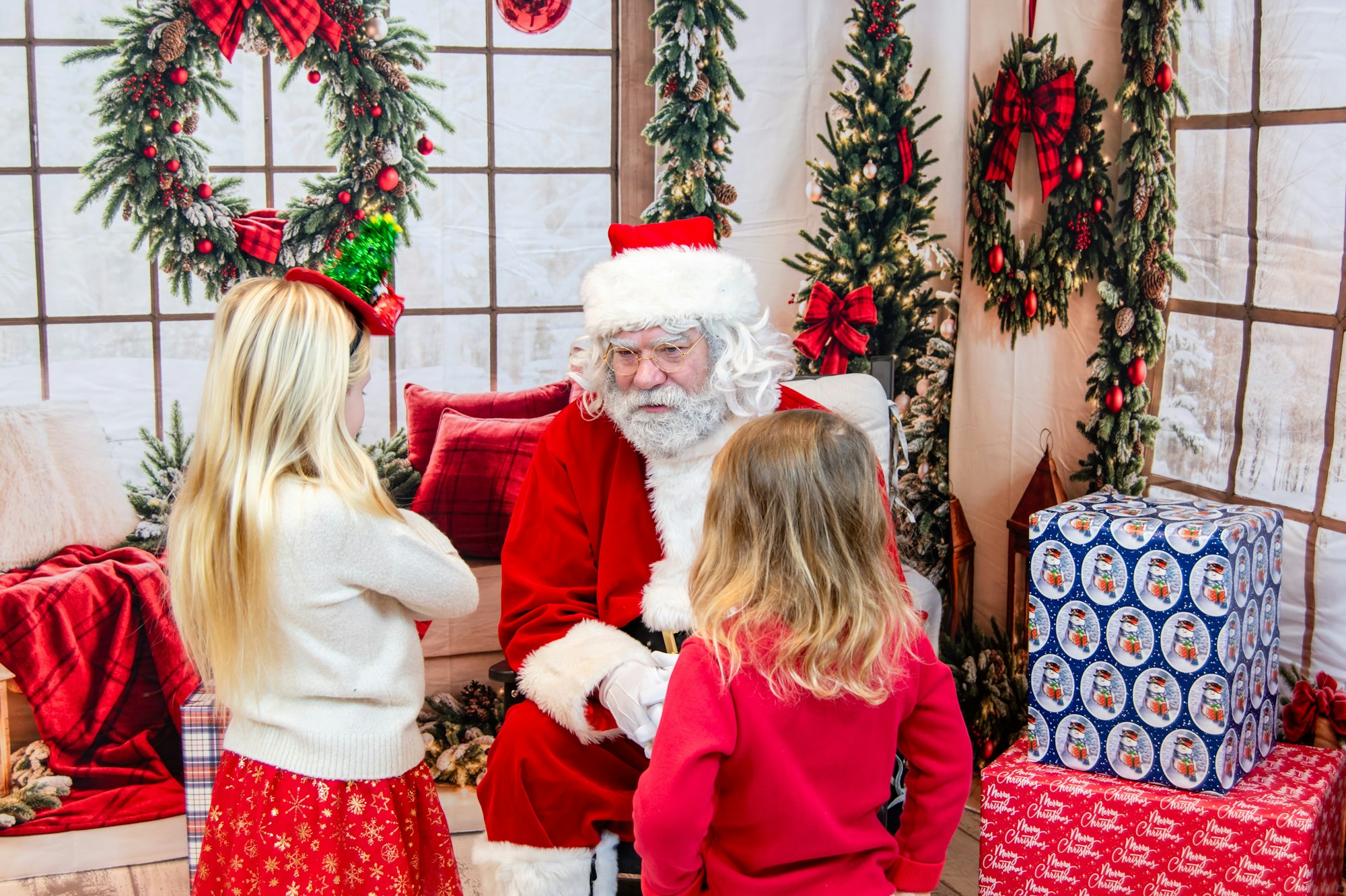Two children talk to Santa Claus in a festive setting with holiday decorations and wrapped gifts around them.