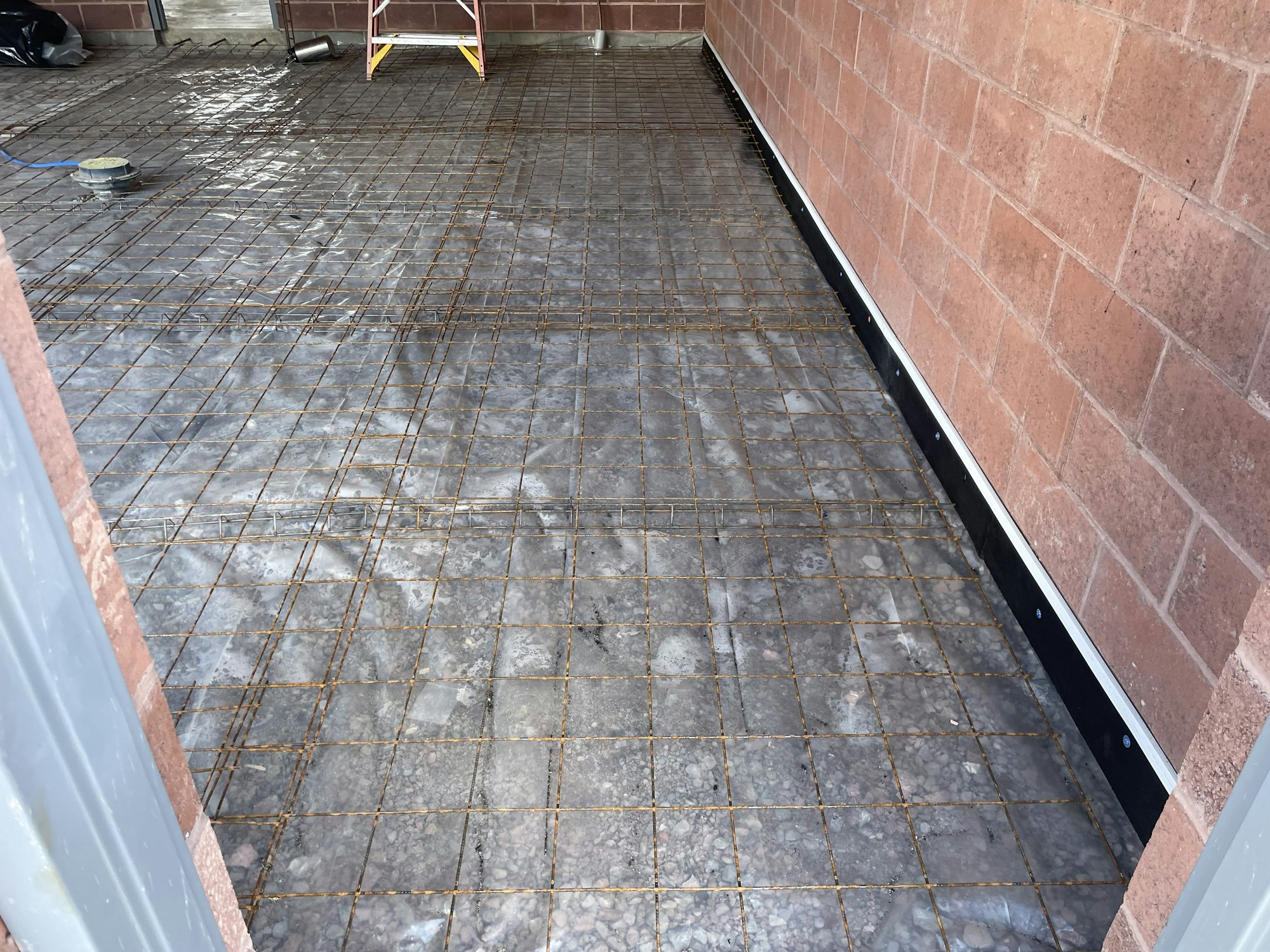 Construction site with rebar grid on floor, preparing for concrete pour, near brick wall and a ladder visible.