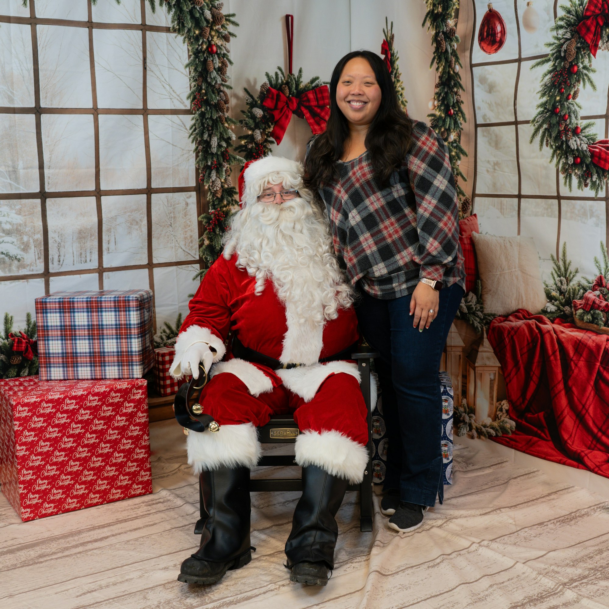 A person poses with Santa Claus in a festive setting with wrapped gifts and holiday decorations.