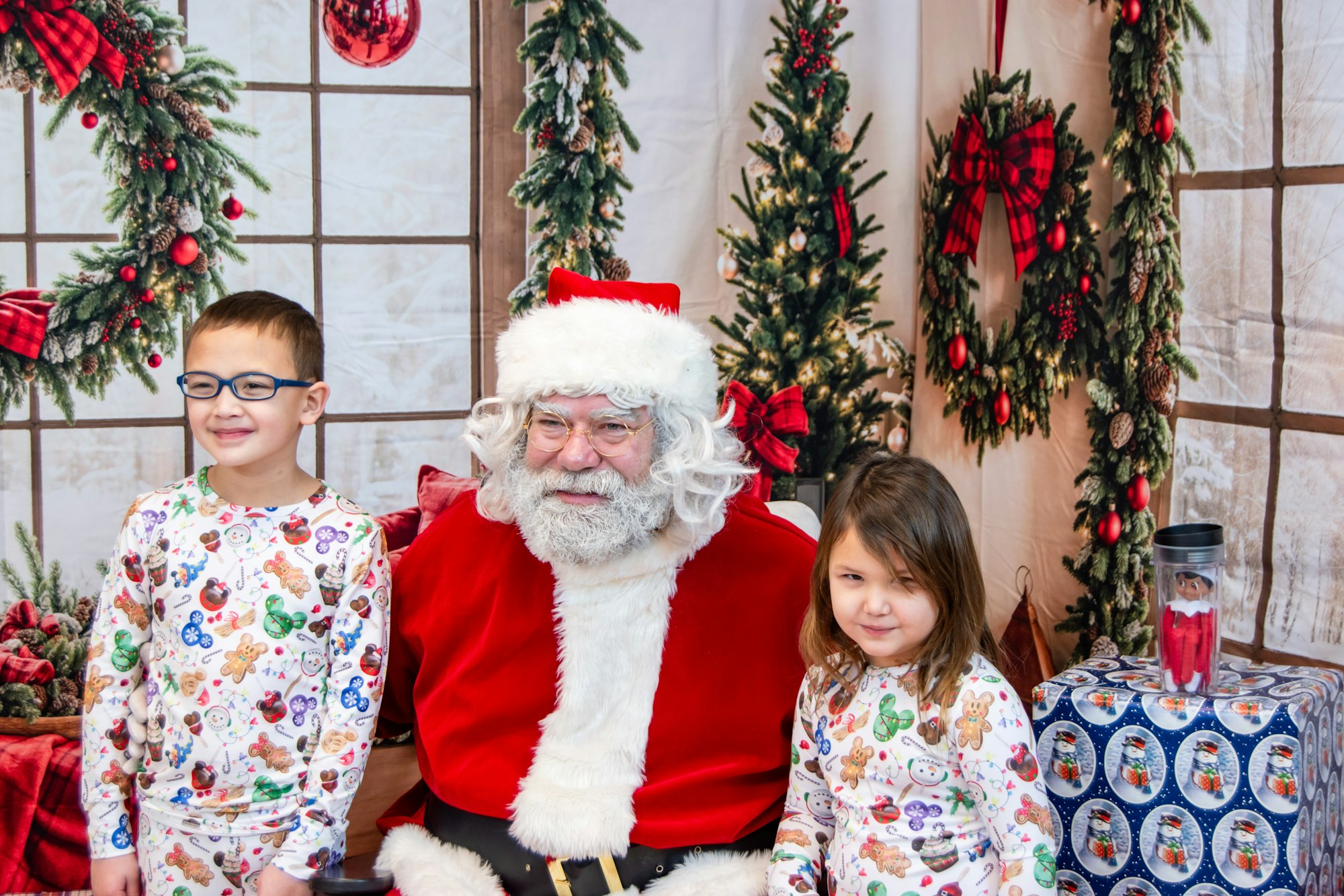 Two children in holiday pajamas sit with Santa Claus, surrounded by festive decorations and a wintery backdrop.