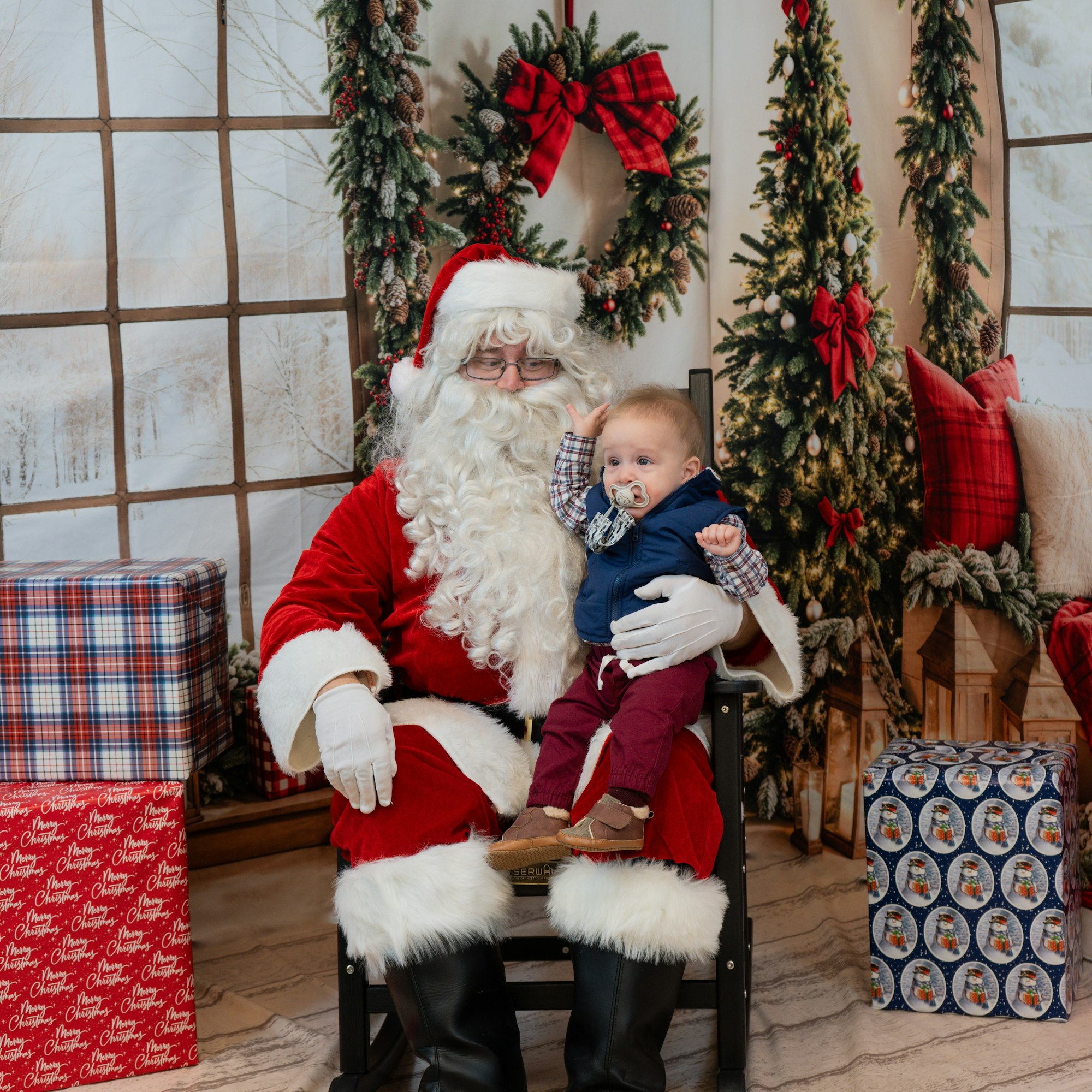 Santa holding a baby with a pacifier, surrounded by Christmas decorations and gifts.