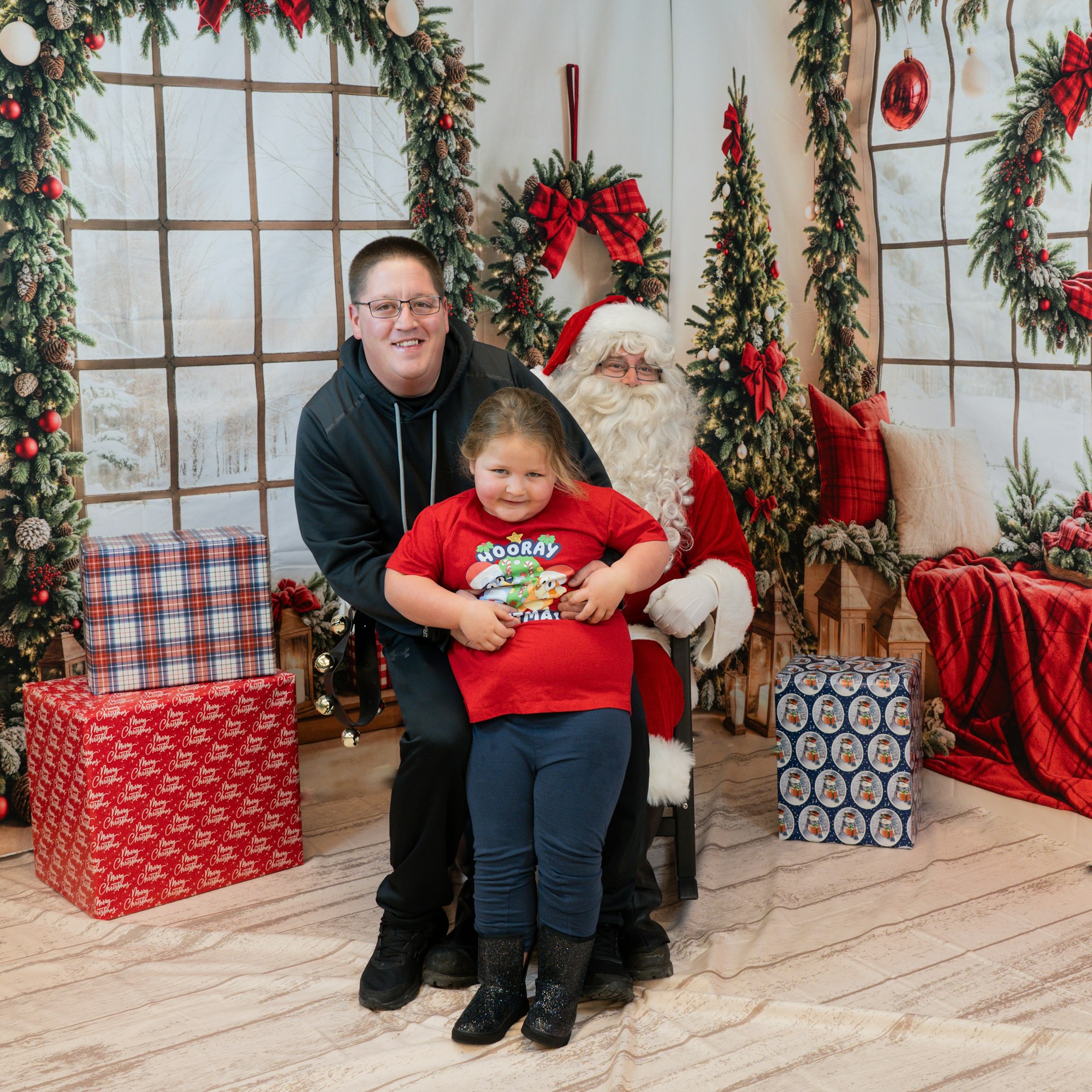 A child and an adult posing with Santa Claus in a festive, holiday setting with wrapped gifts and decorations.