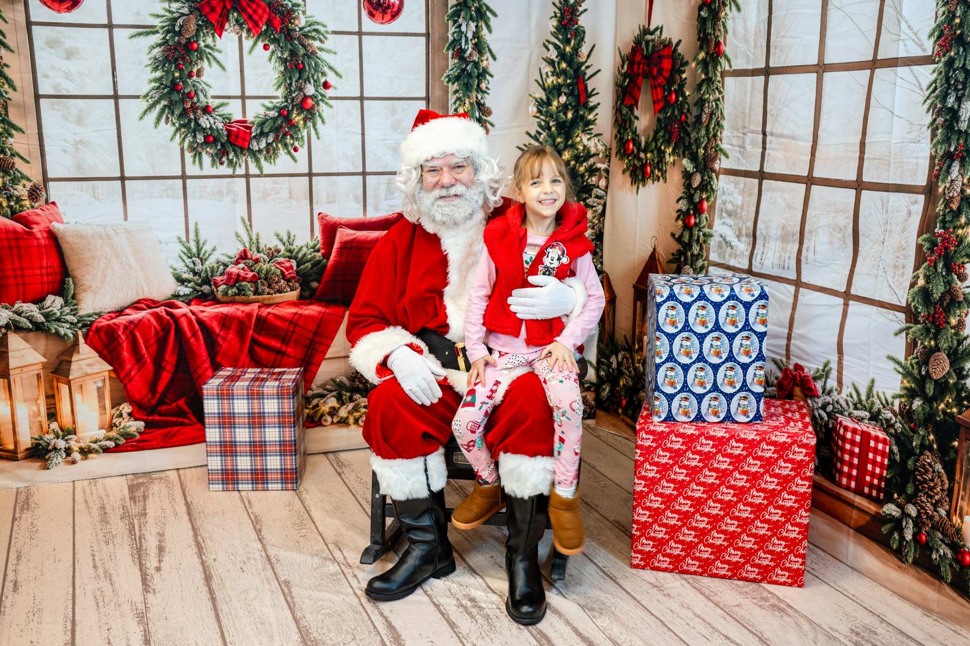 A child sits on Santa’s lap in a festive setting with Christmas decorations and wrapped gifts around them.