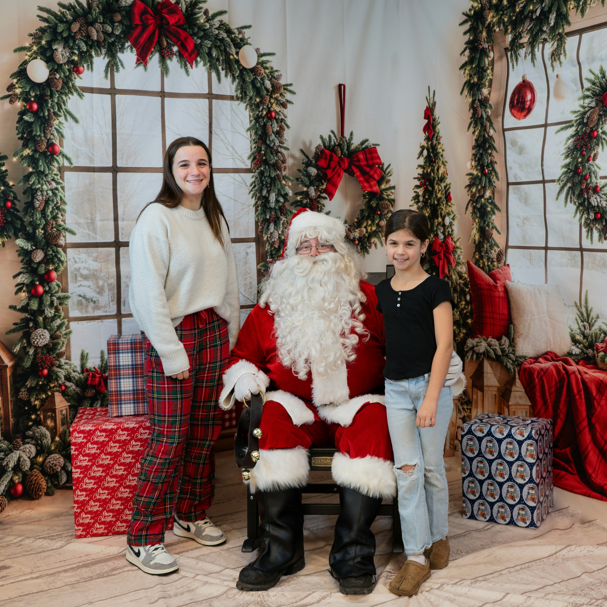 Two people pose with Santa in a festive setting with holiday decorations and wrapped gifts.