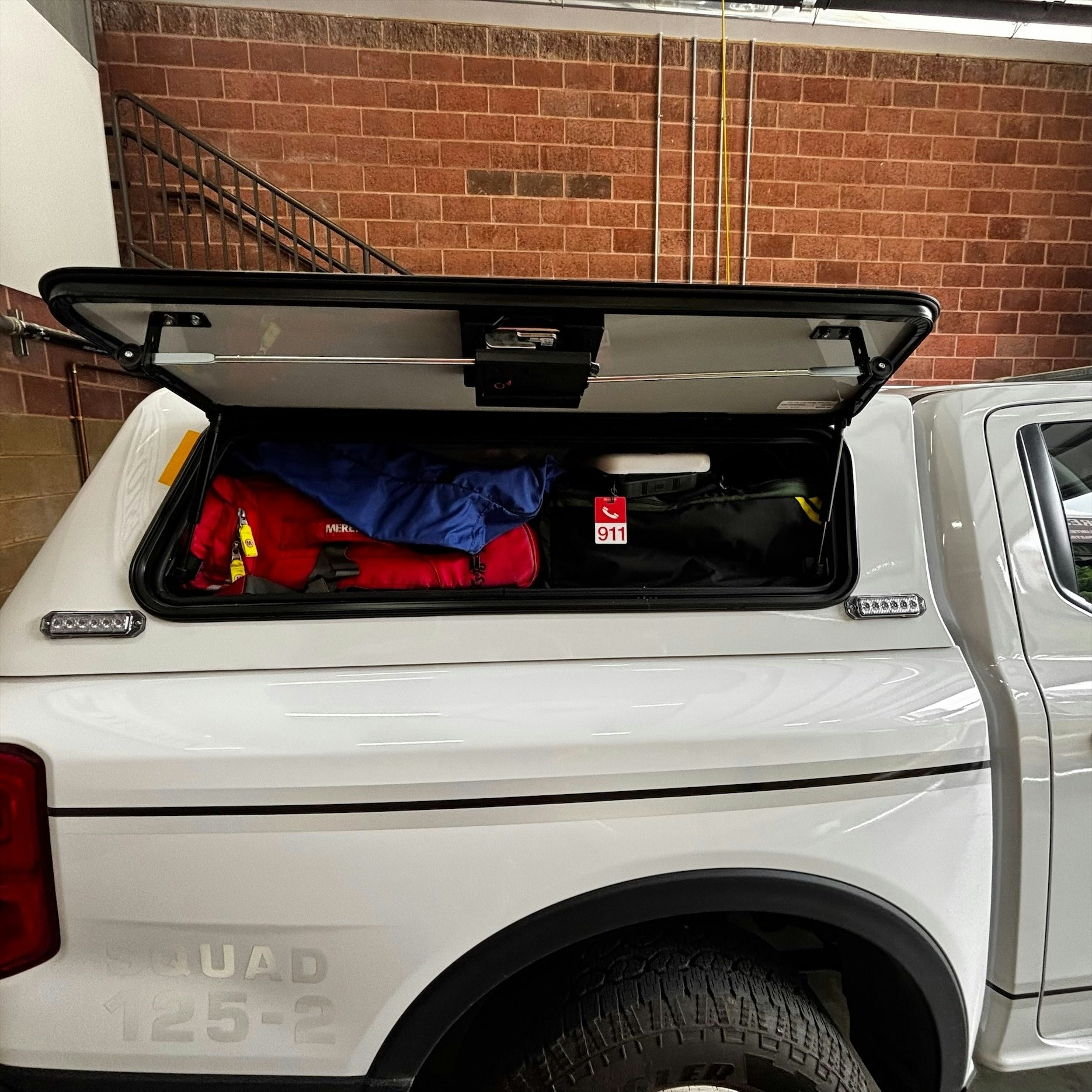 The image shows a truck bed cover open, revealing various bags and equipment, including a red emergency bag marked "911."
