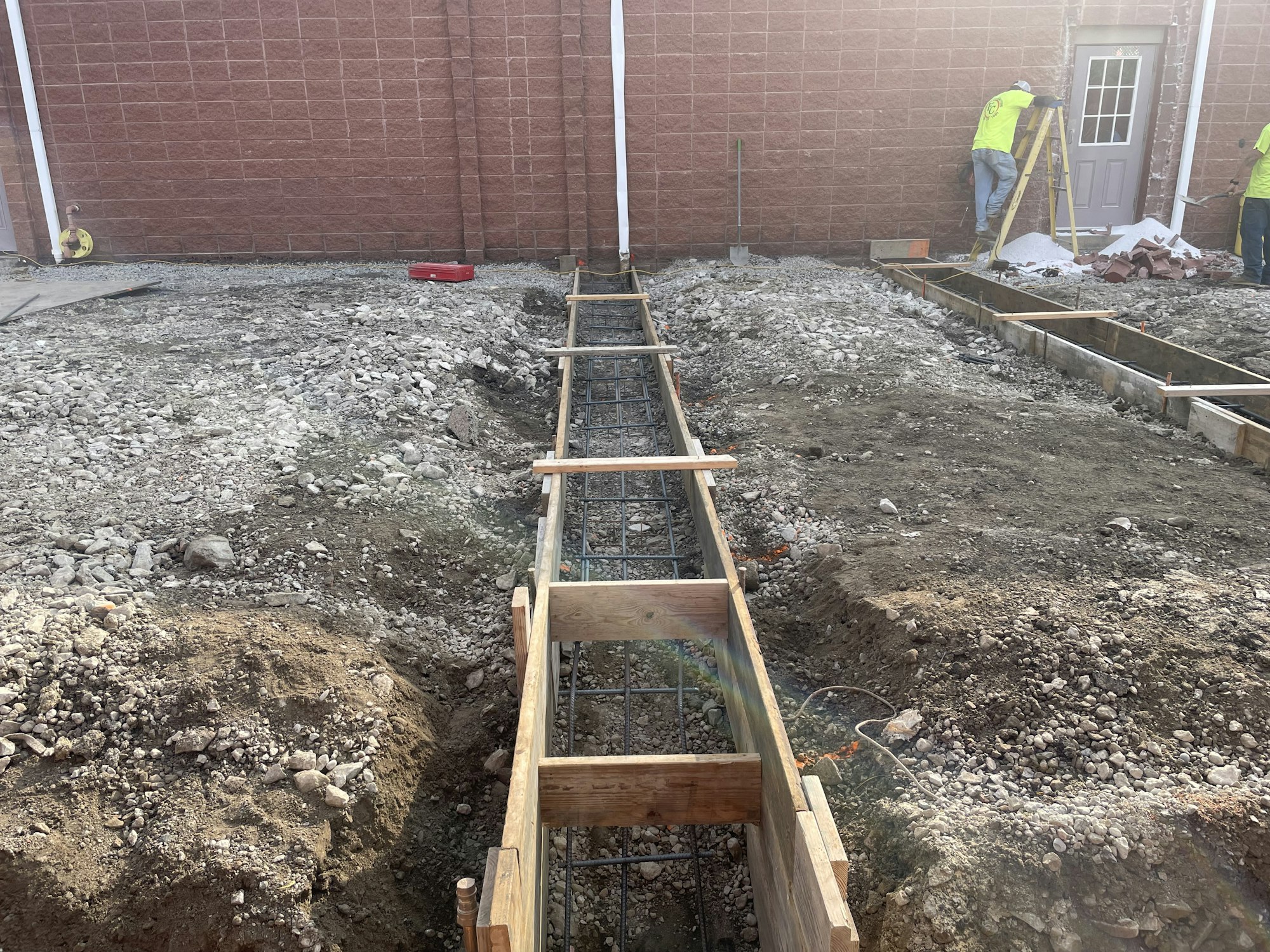 Construction site with workers, trench, wooden forms for concrete, and building in background.