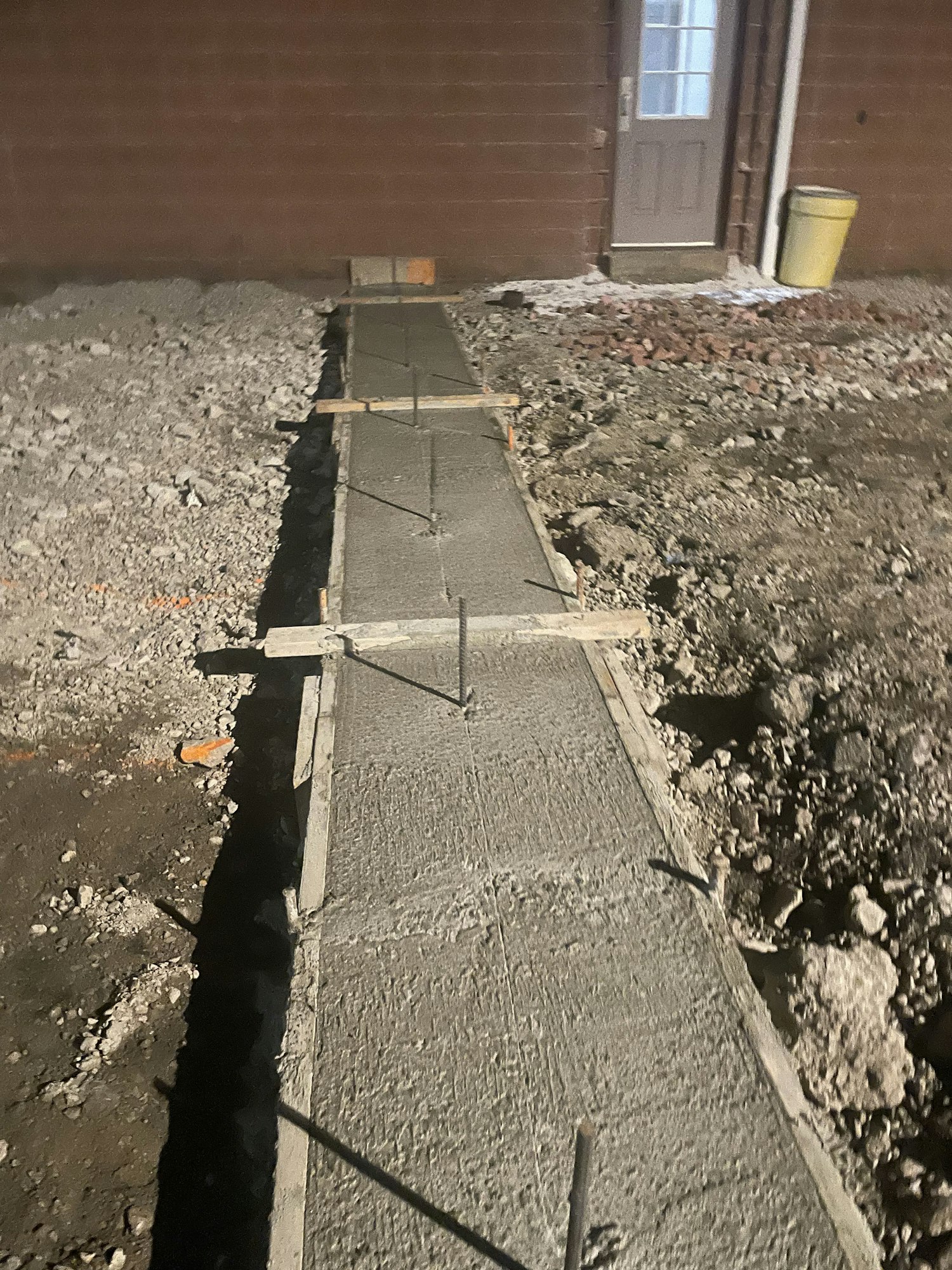 Freshly poured concrete foundation wall with wooden formwork still attached, near a building.