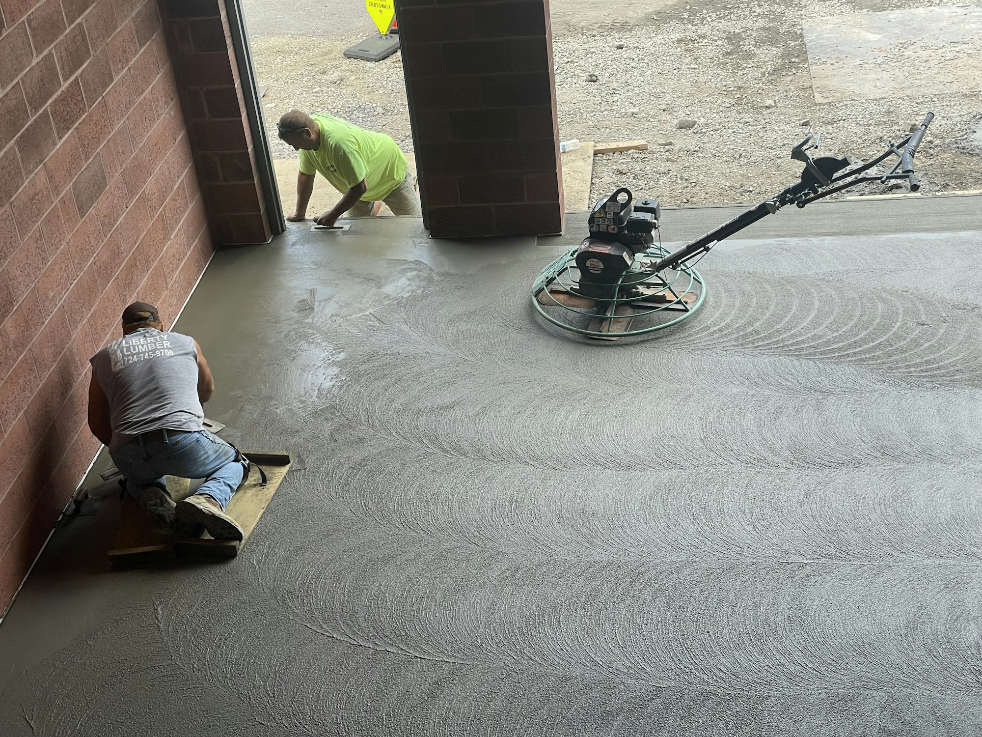 Two men working on a concrete floor, one using a float machine, and the other hand-finishing edges.