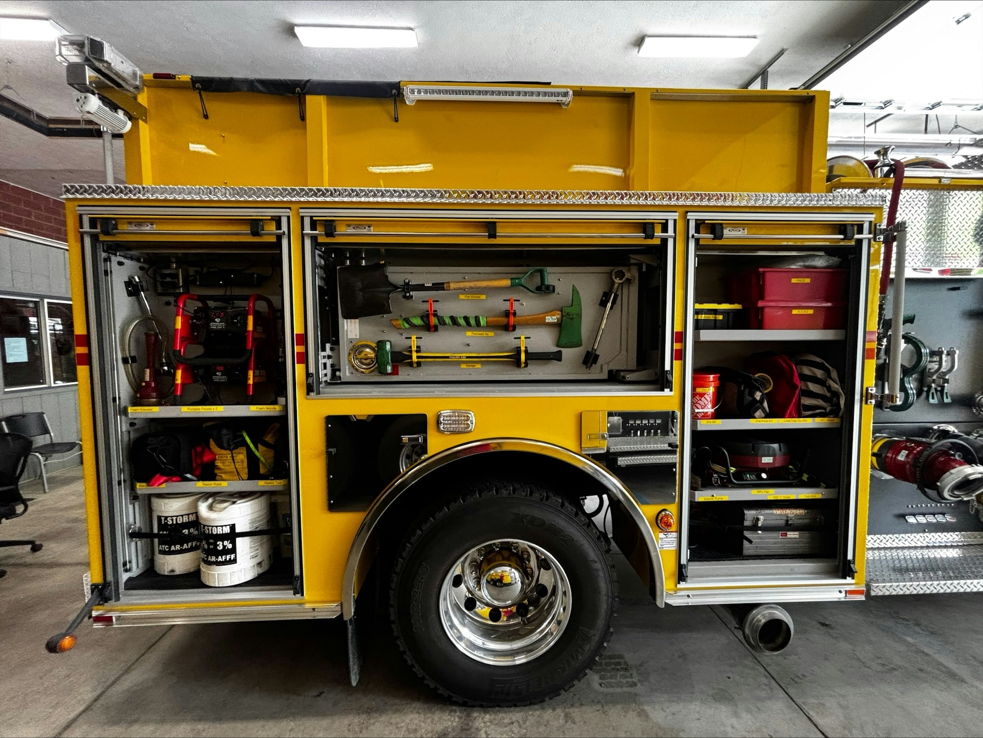 An organized side of a yellow firetruck features tools, equipment, hoses, and firefighting supplies neatly stored in compartments.