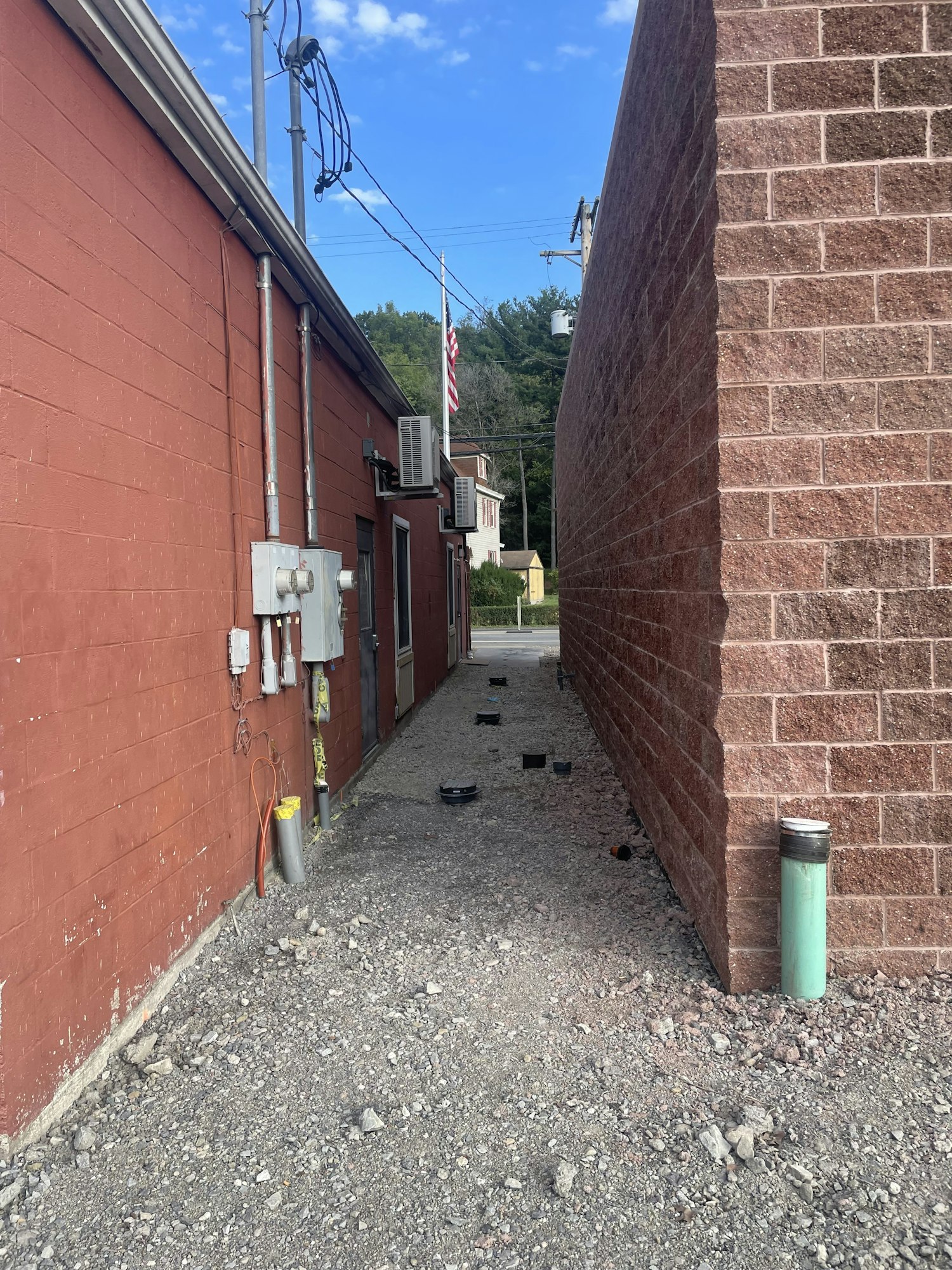Side alley with utility pipes on a wall, gravel ground, and red building, image is rotated 90 degrees clockwise.