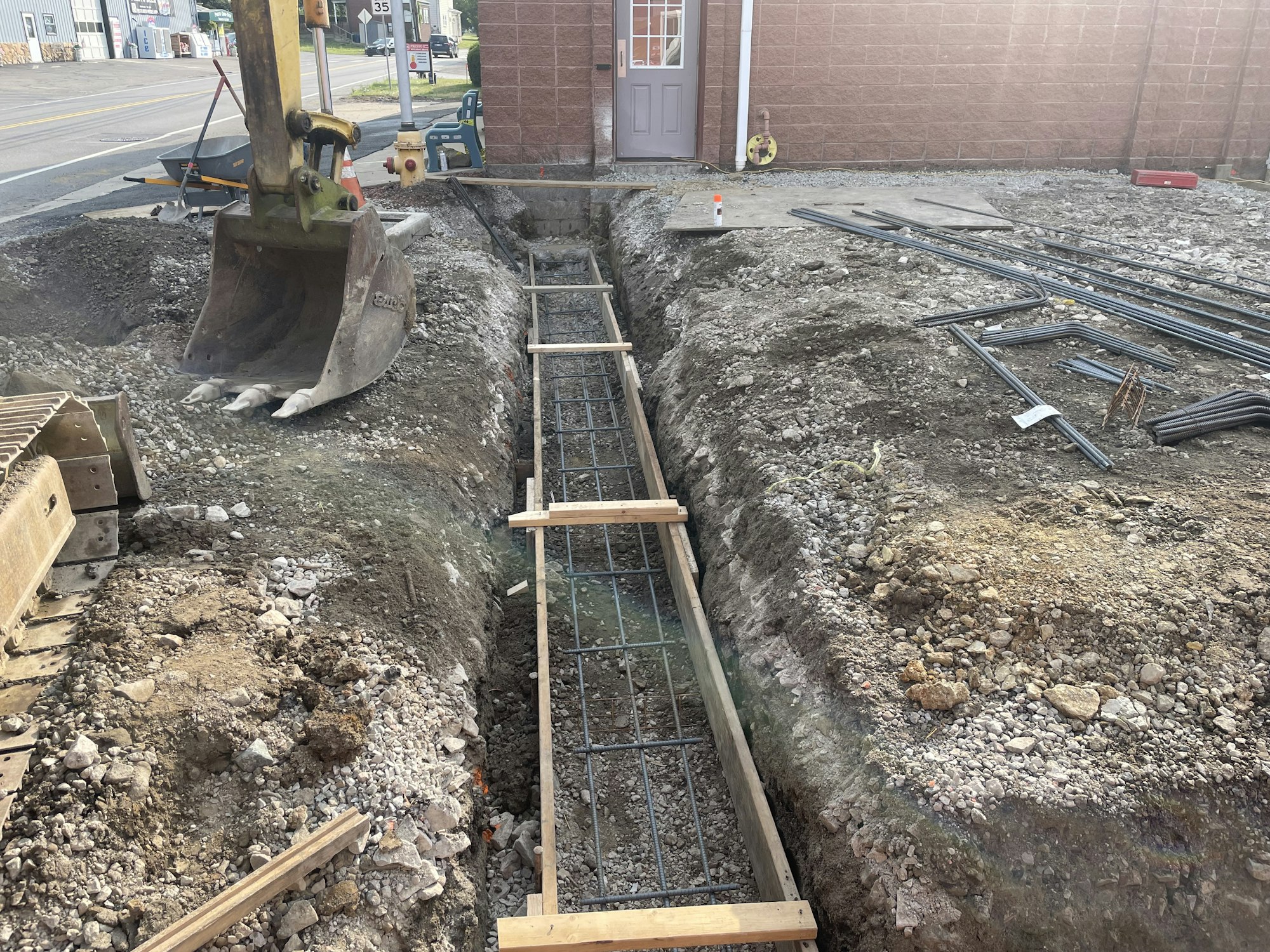 Construction site with excavator, trench, rebar, and wooden bracing for concrete work.