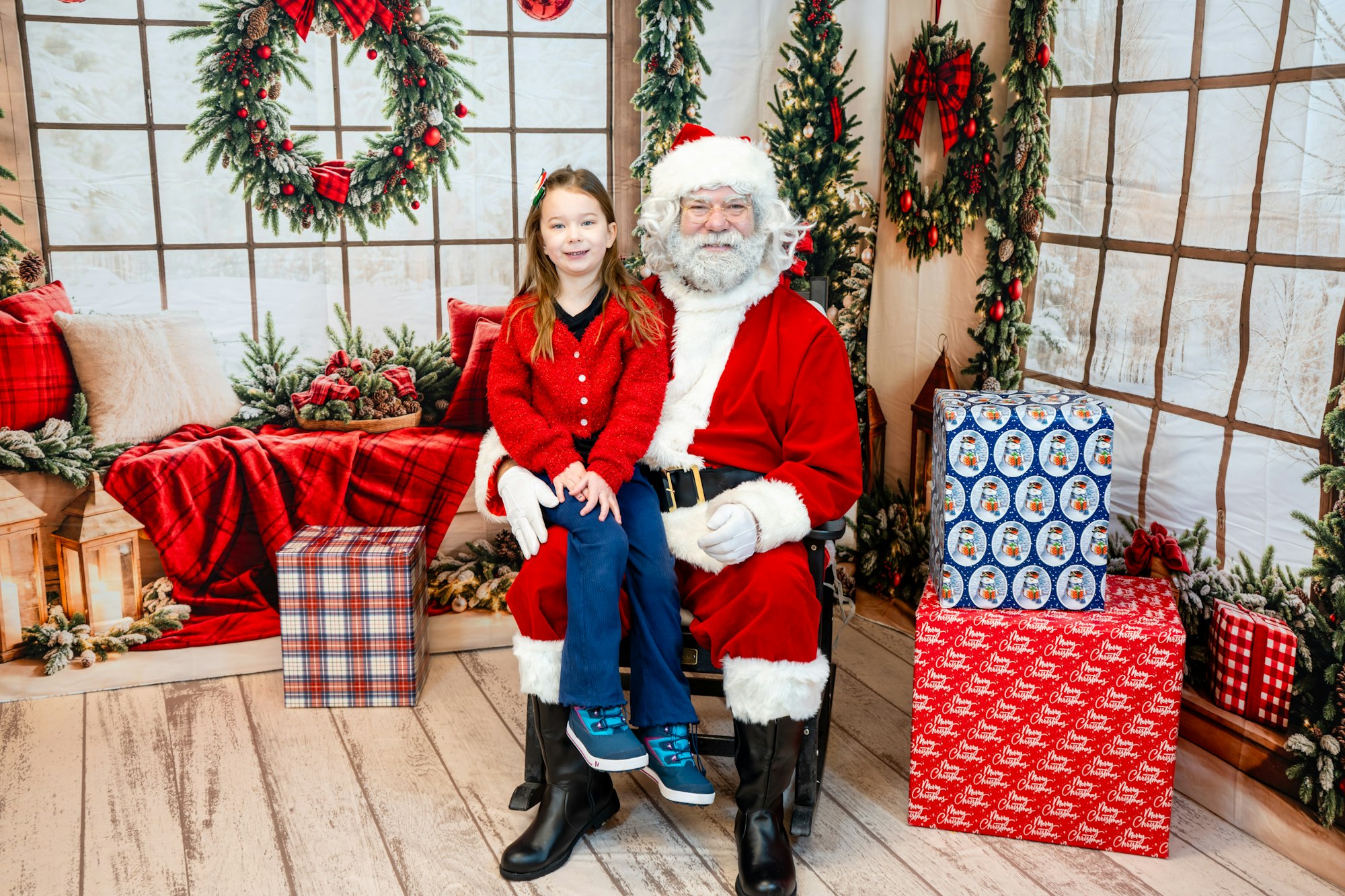 A child in a red sweater sits on Santa's lap in a festive setting with holiday decorations and wrapped gifts around them.