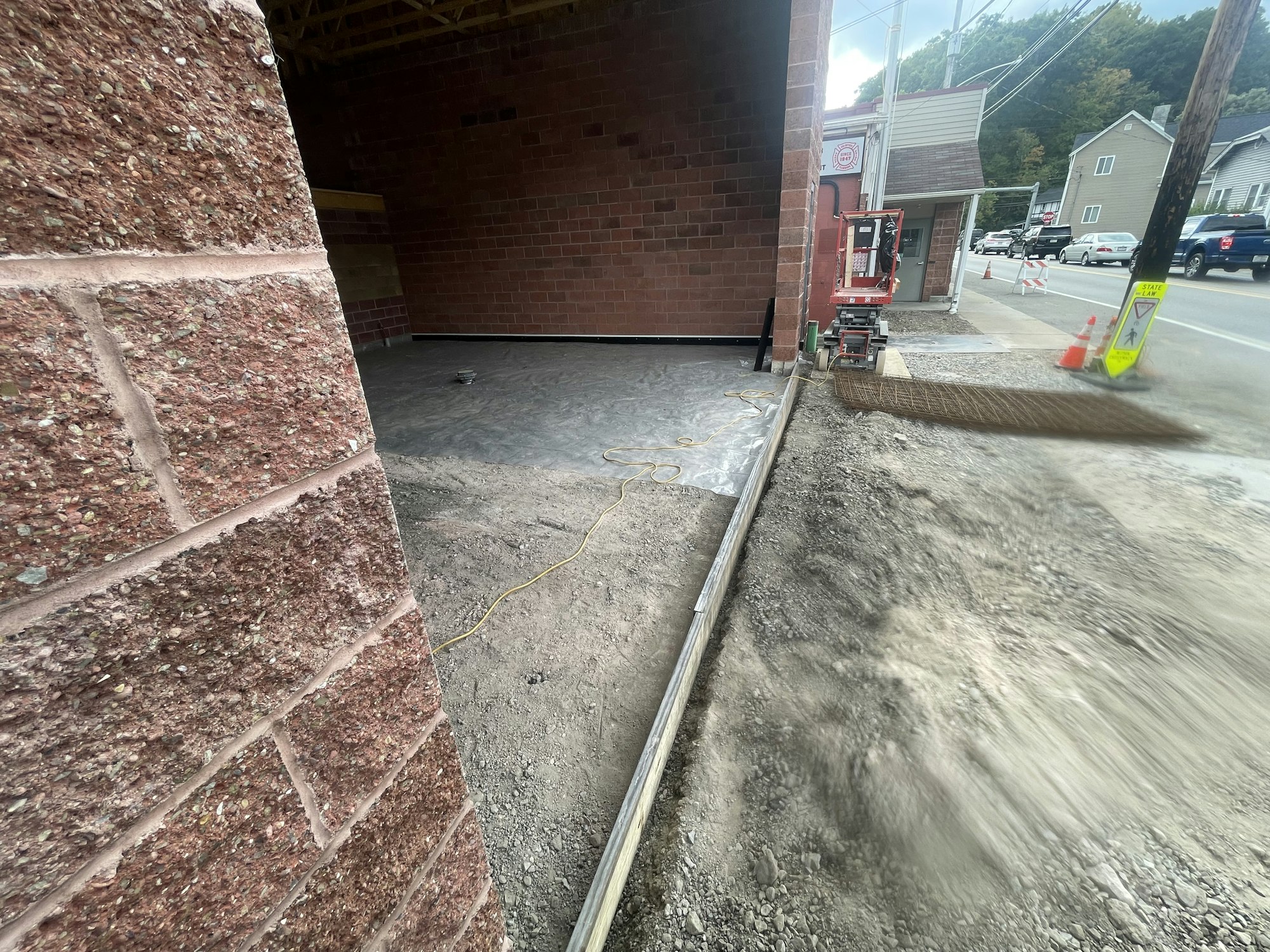 Fresh concrete floor next to a gravel area, with construction equipment and a road with cars and traffic cones visible.