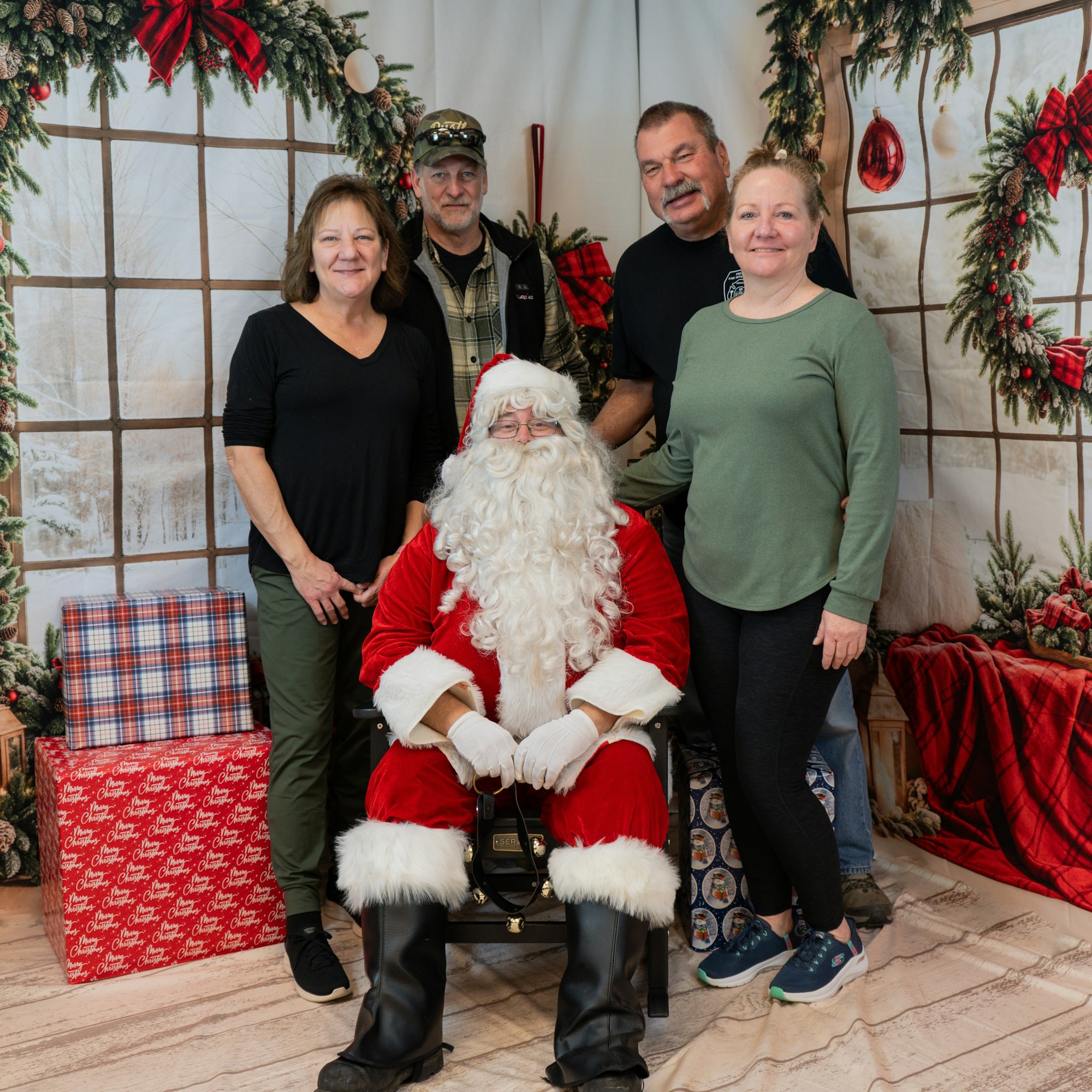 A group photo with four adults and Santa Claus seated, surrounded by Christmas decorations and gifts.