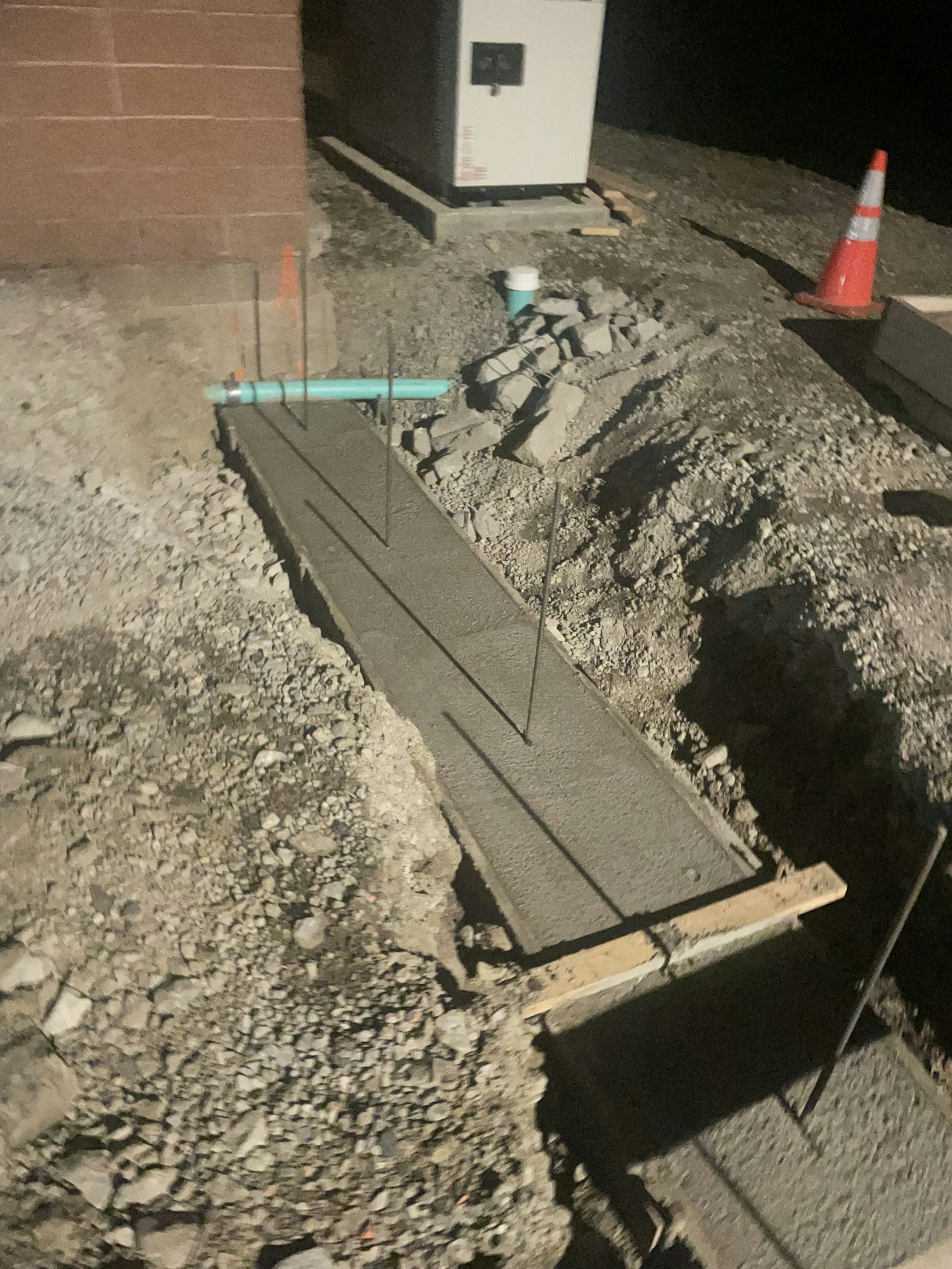 A construction site with exposed pipes, a concrete formwork, rubble, and a safety cone at night.