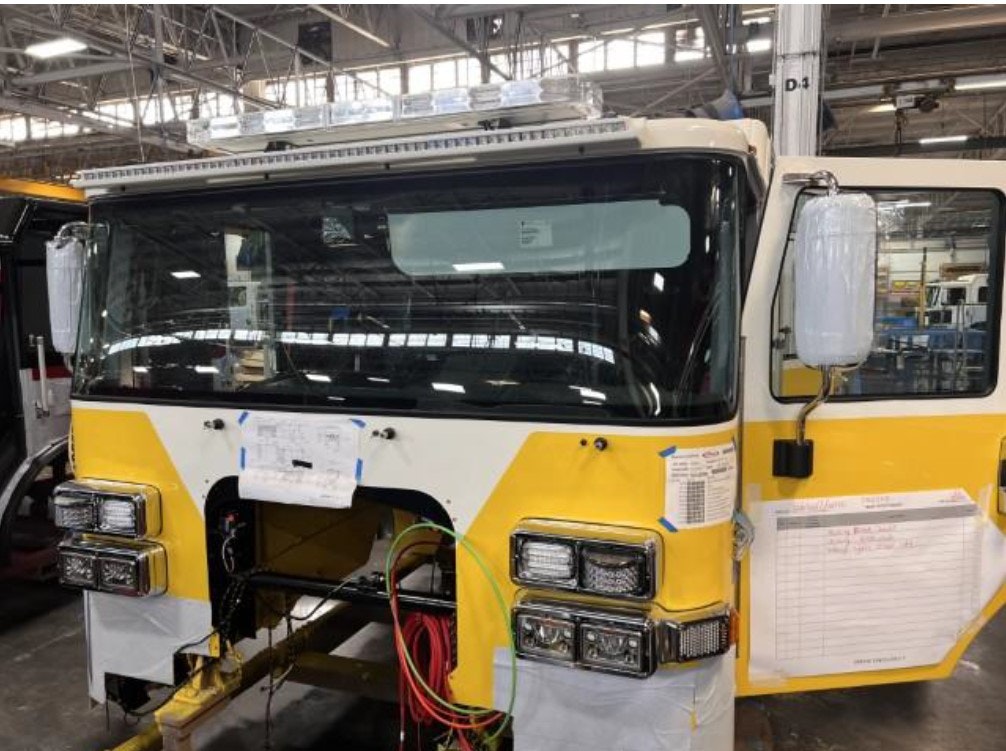 Yellow and white fire truck cab in a factory, partially assembled with notes and lists attached.