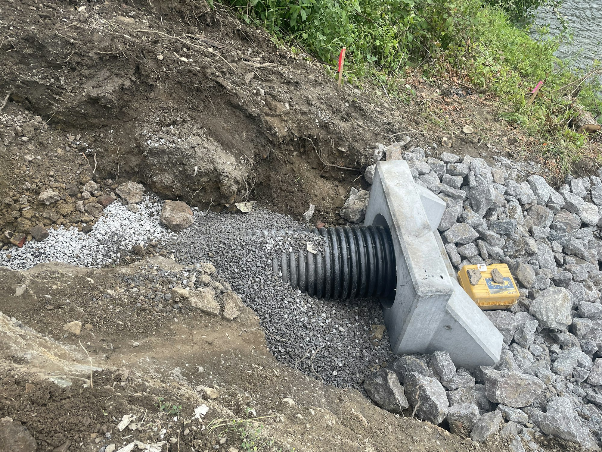 A drainage culvert being installed near a water body with rocks, gravel, and excavation visible.