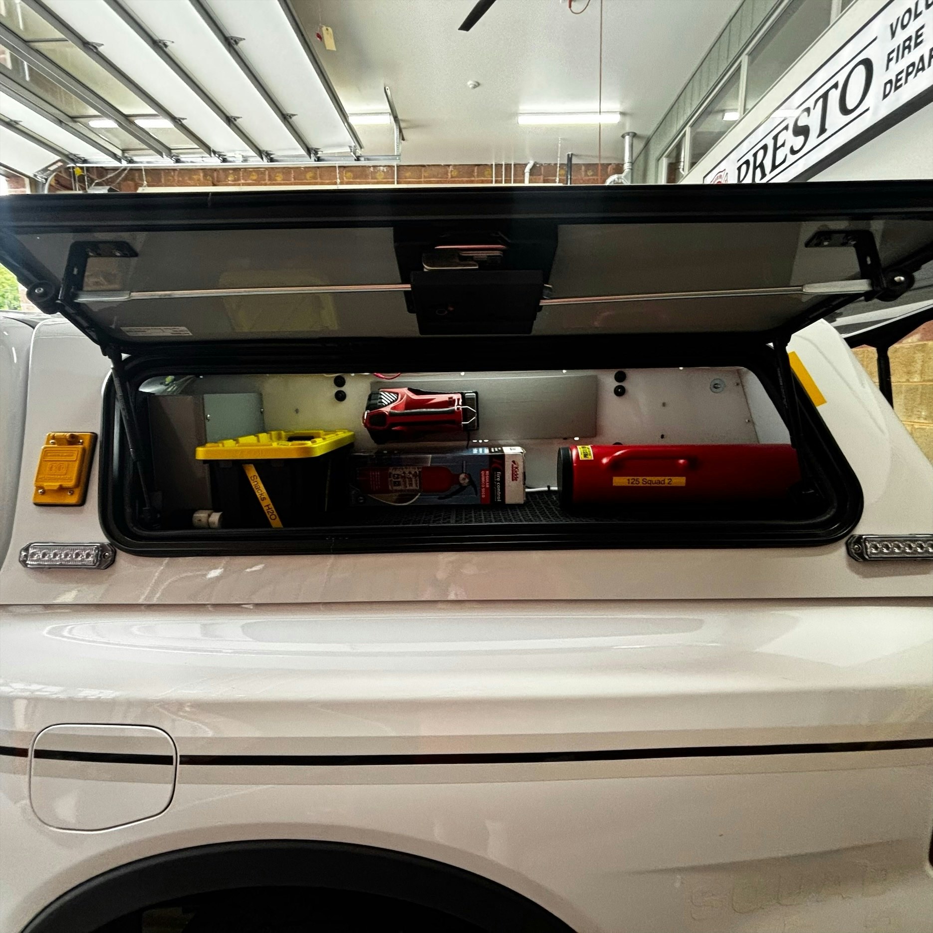 The image shows an open truck cap with storage containing tools, a fire extinguisher, and a yellow container inside.