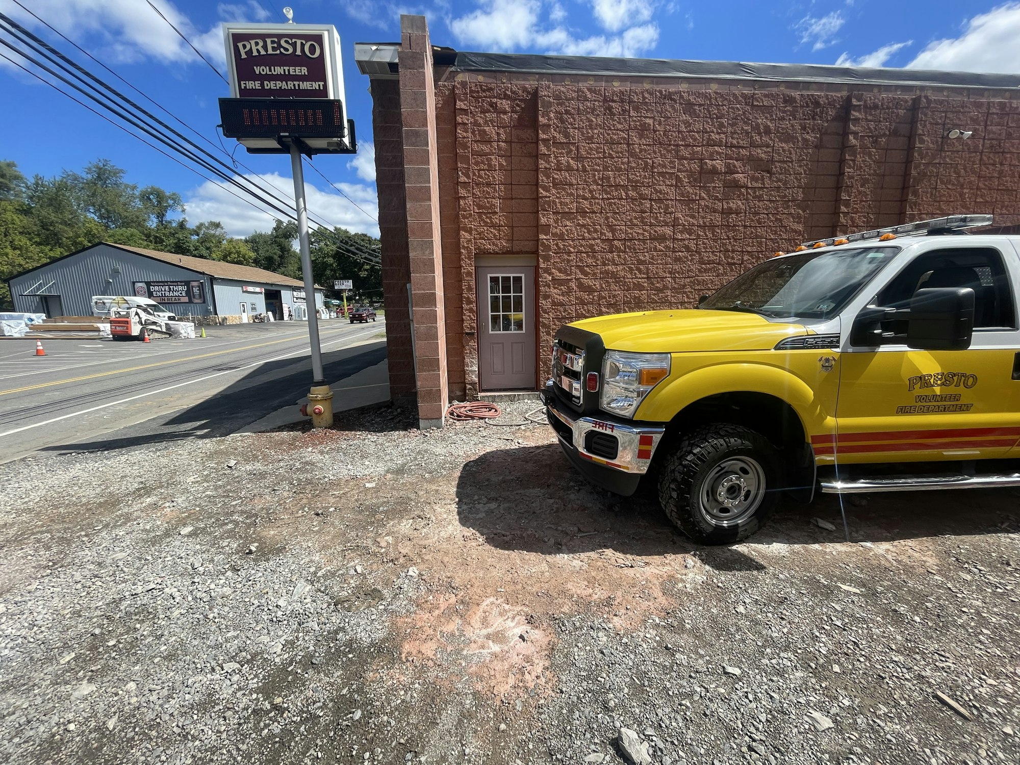 A yellow fire department truck near a sign reading "Presto Volunteer Fire Department," with a building and road in the background.