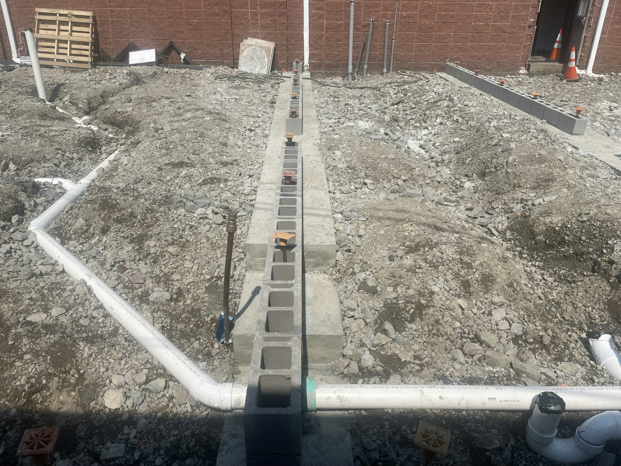 A construction site with exposed plumbing and cinder blocks, with rocky soil and building in the background.