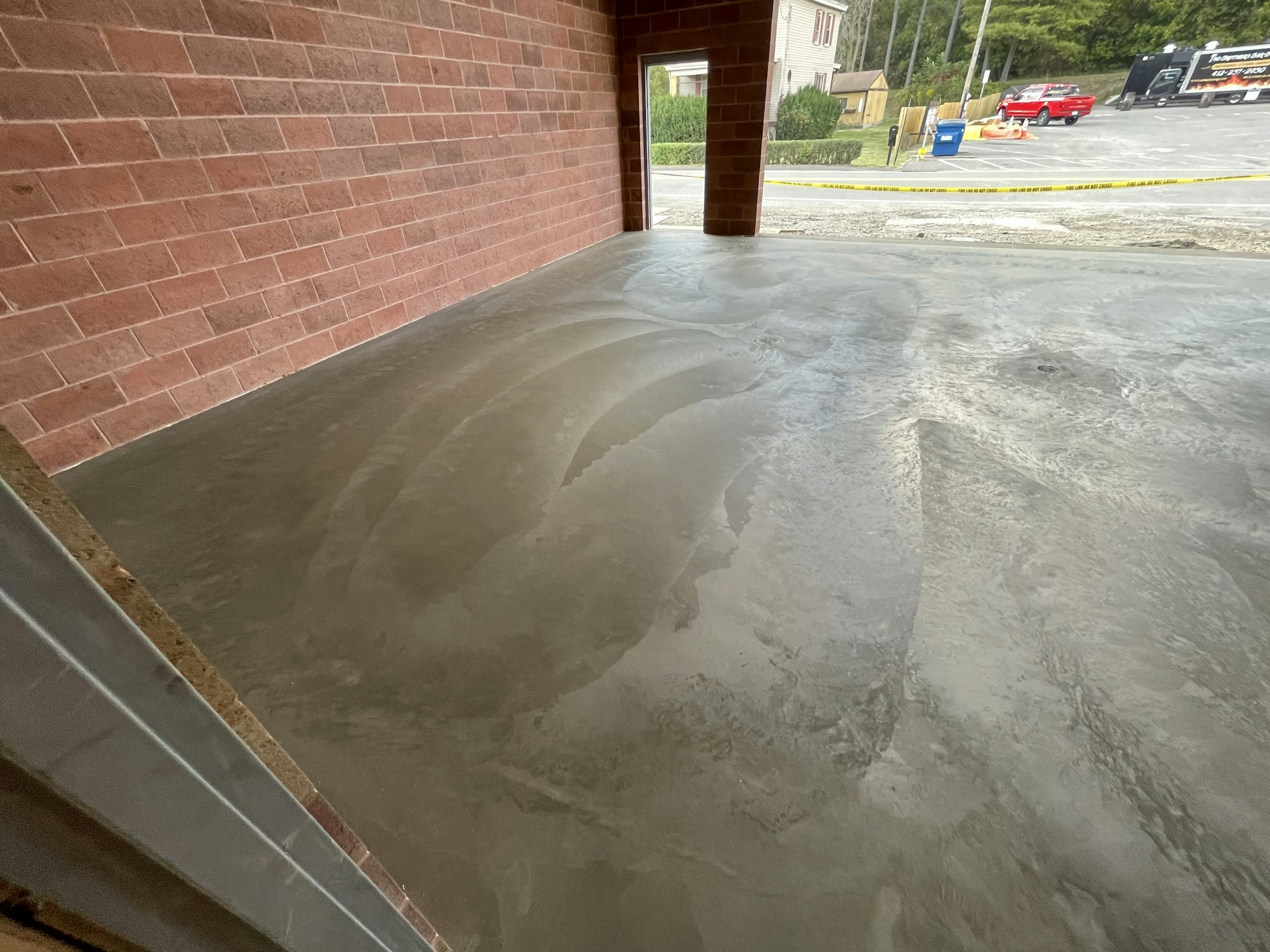 A wet concrete floor with tire track impressions, brick wall to the left, and a parking lot seen through an open garage door.