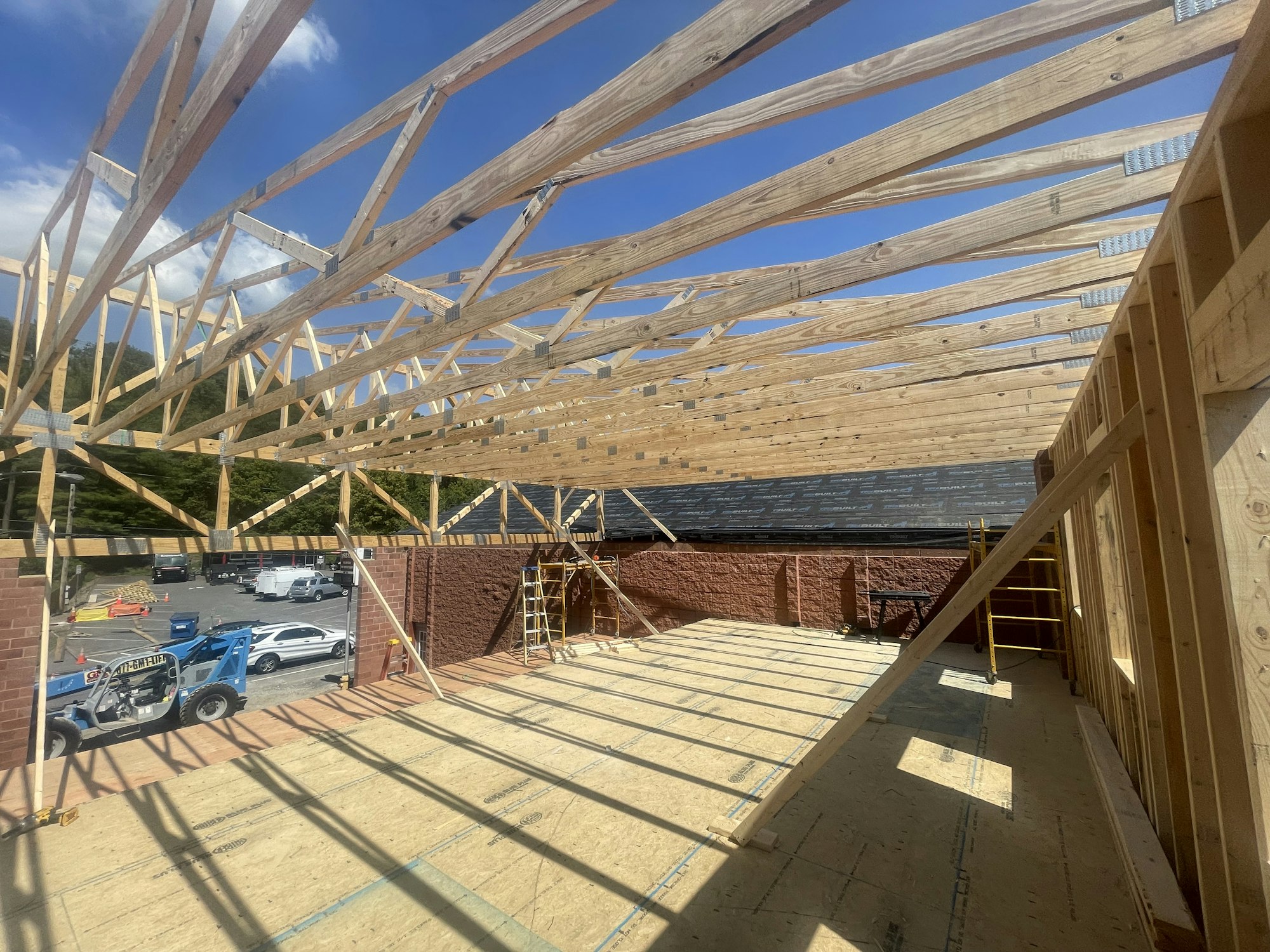 Construction site with wooden roof trusses, a plywood-covered floor, and a telehandler vehicle outside.