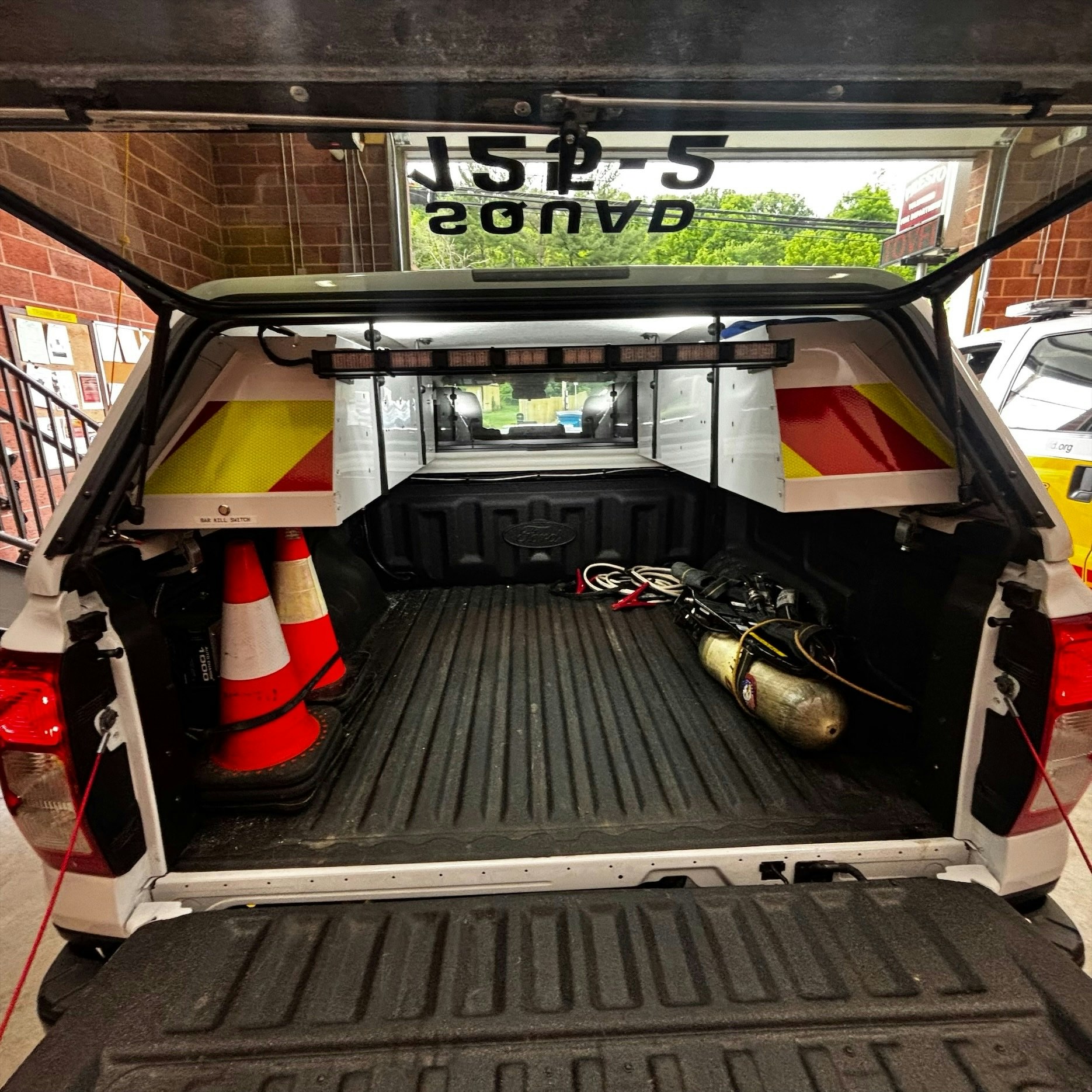 The image shows the open back of a vehicle with safety cones, equipment, and storage compartments, likely used for emergency services.