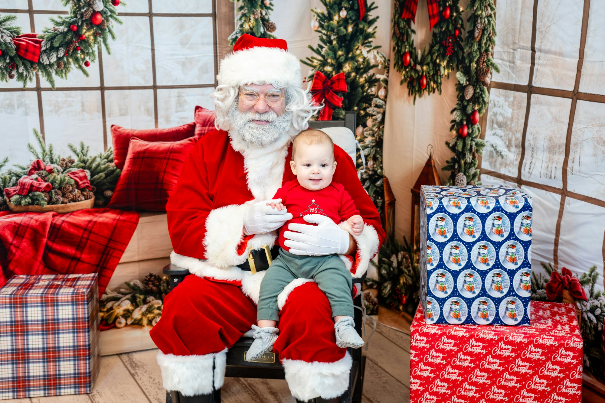A jolly Santa Claus sits in a festive setting with a smiling baby, surrounded by Christmas decorations and gift boxes.