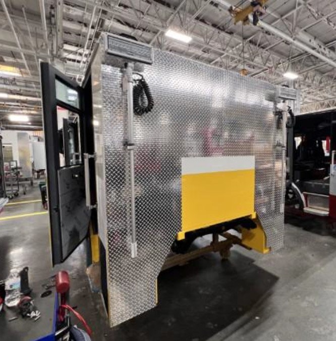 A shiny metal truck or vehicle component with a yellow section, in an industrial setting with open doors and coiled cables.